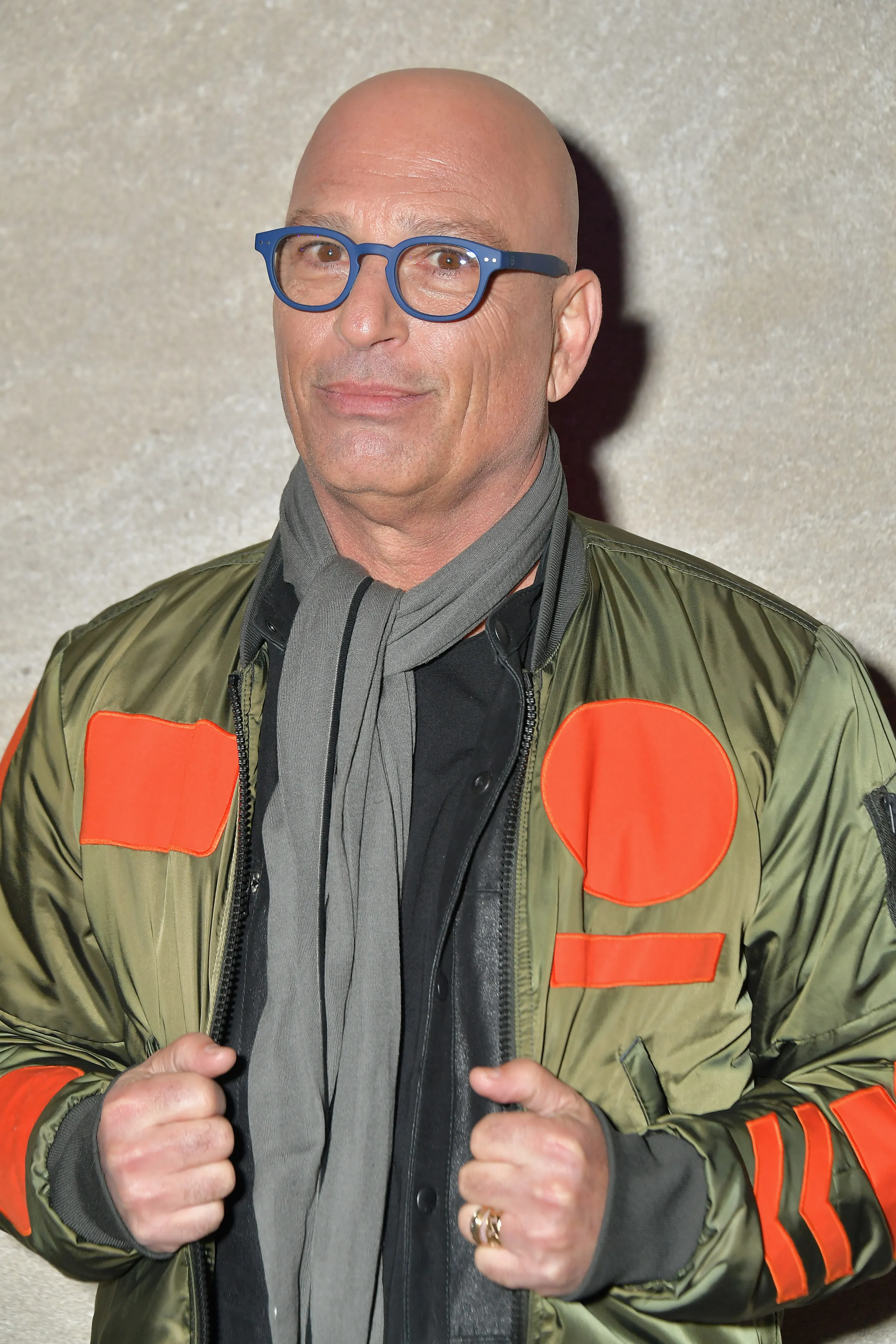 Howie Mandel wearing a green bomber jacket with orange patches, a gray scarf, and blue-framed glasses.