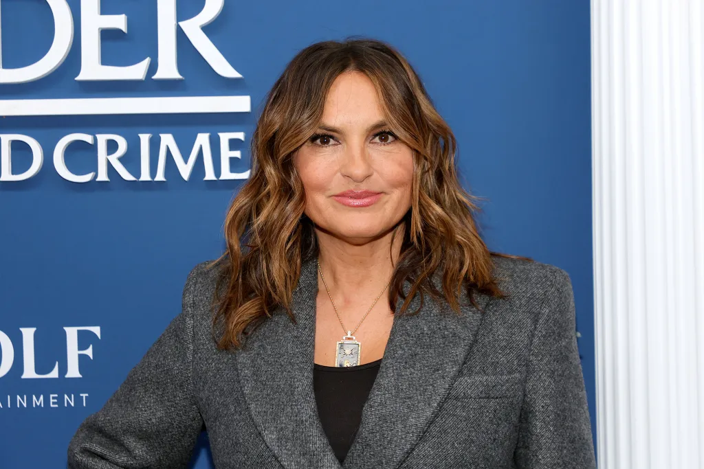 Mariska Hargitay smiling in a grey blazer with a black top and a rectangular pendant necklace.