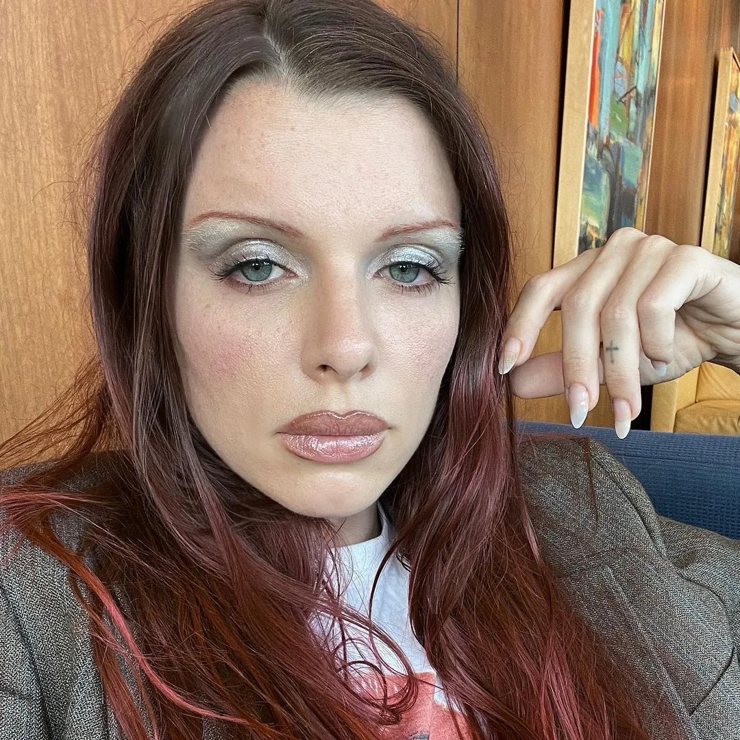 Julia Fox with red hair, frosted eyeshadow, and a cross tattoo on her finger.