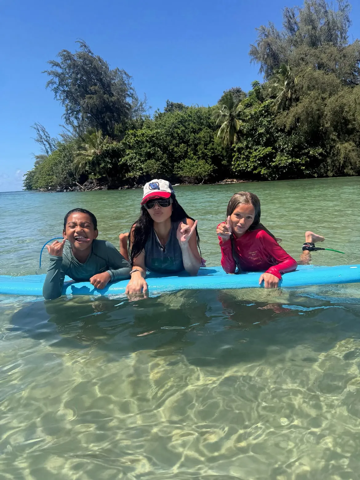 Kim Kardashian and two children posing on a surfboard in clear ocean water.