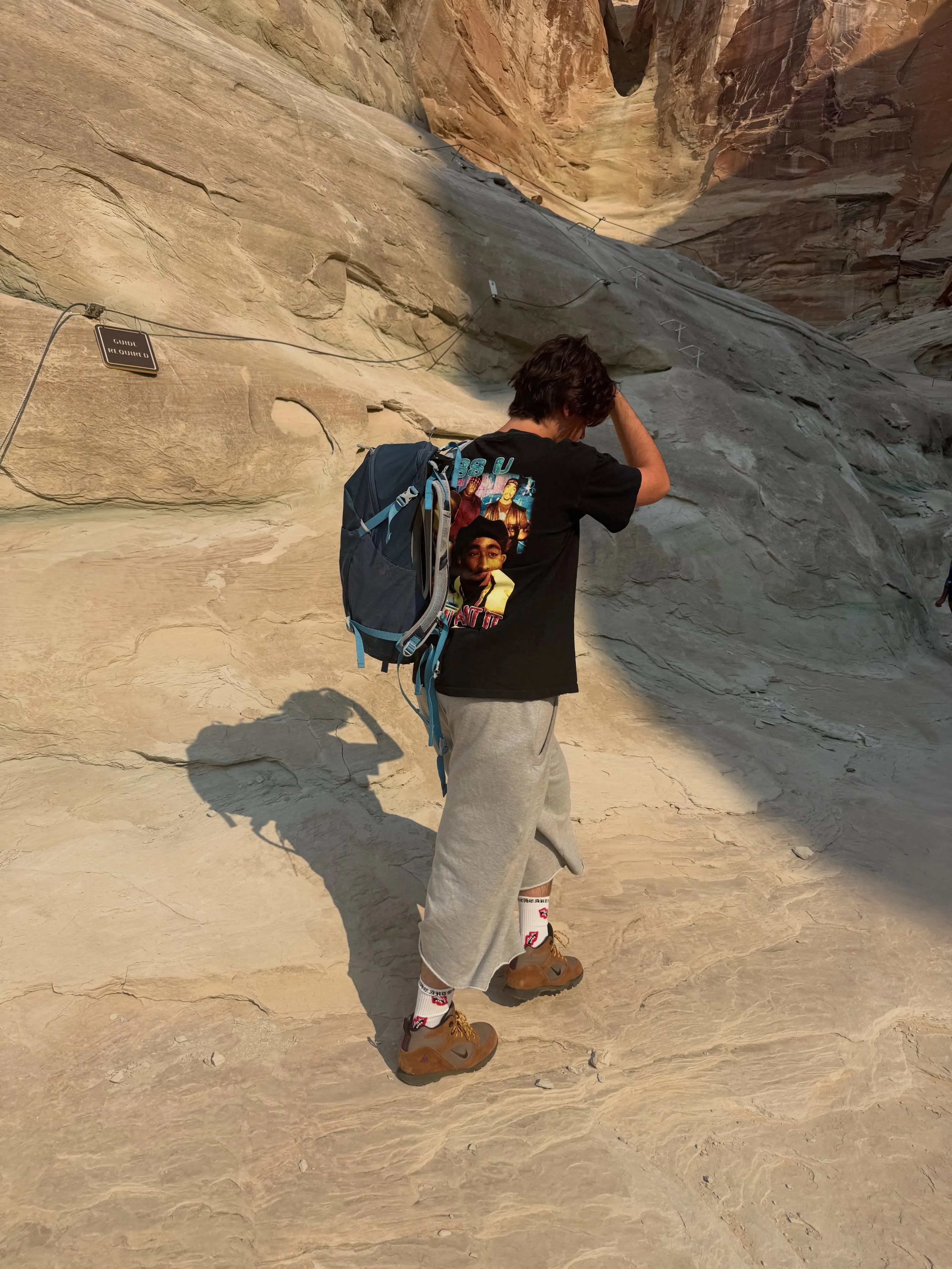 Mason Disick with a backpack hiking up a steep, rocky path with a "GUIDE REQUIRED" sign visible.
