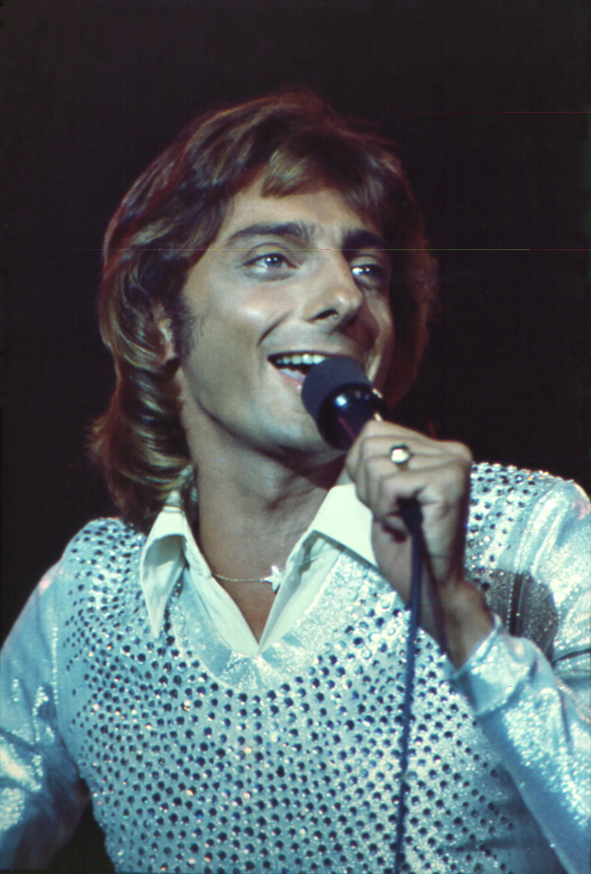Barry Manilow singing into a microphone, wearing a shiny shirt with a star necklace.
