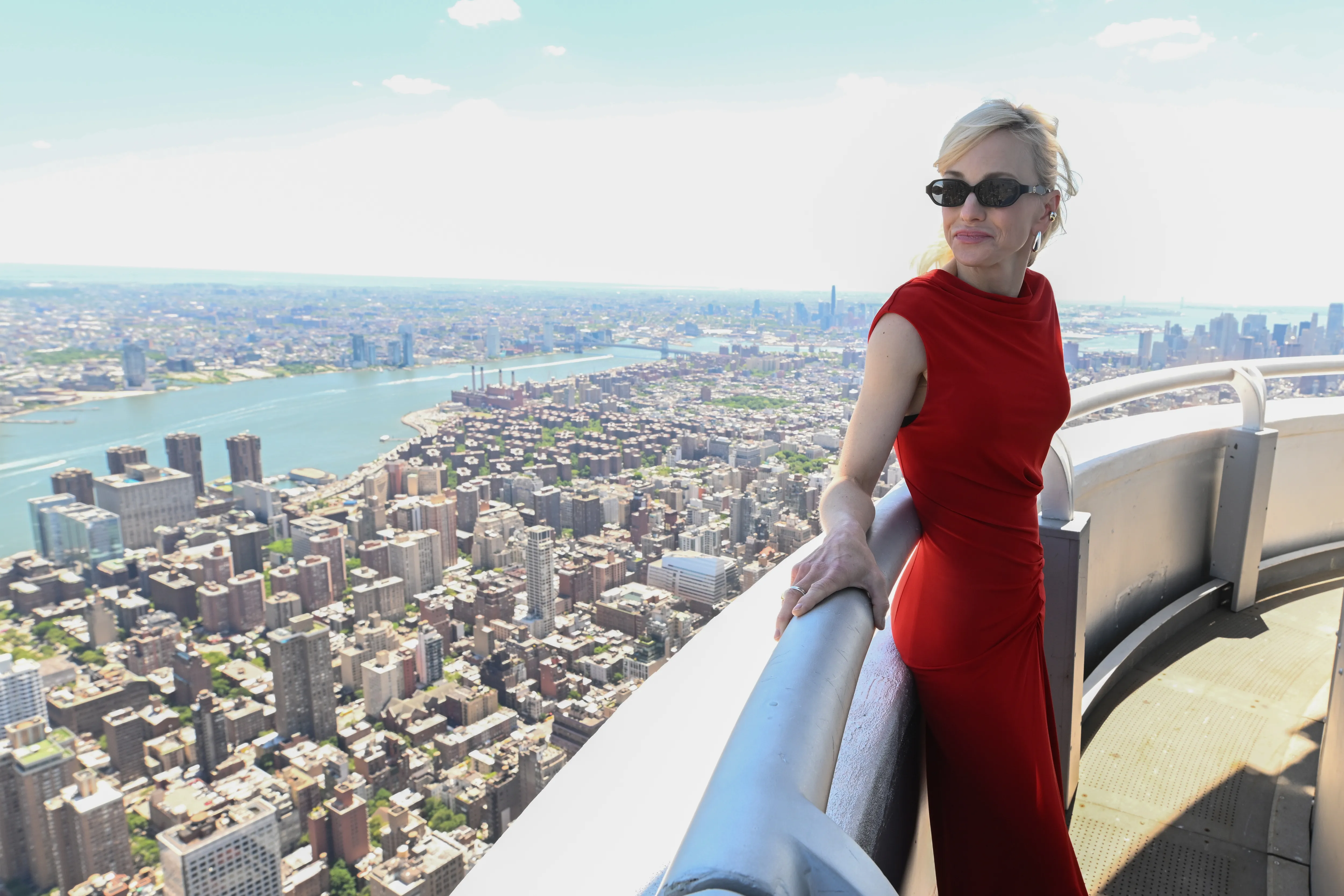 Anna Faris in a red dress and sunglasses on the observation deck of the Empire State Building, overlooking New York City.