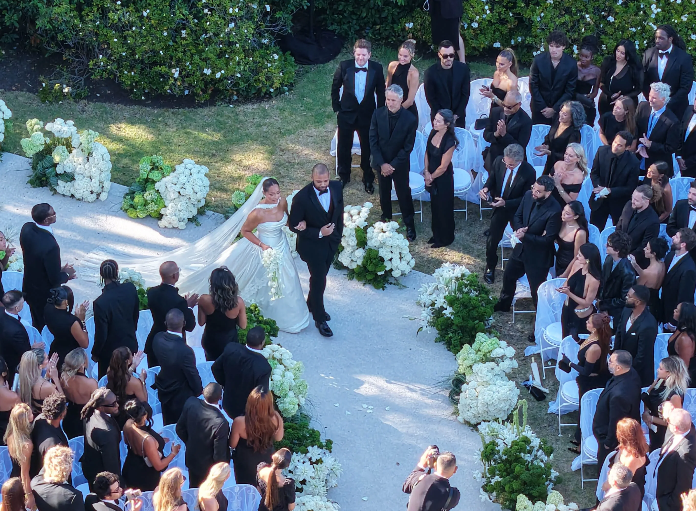 Jordyn Woods in a white wedding gown walks arm-in-arm with a man in a black tuxedo down an aisle lined with white flowers, while guests in black attire watch from white chairs.