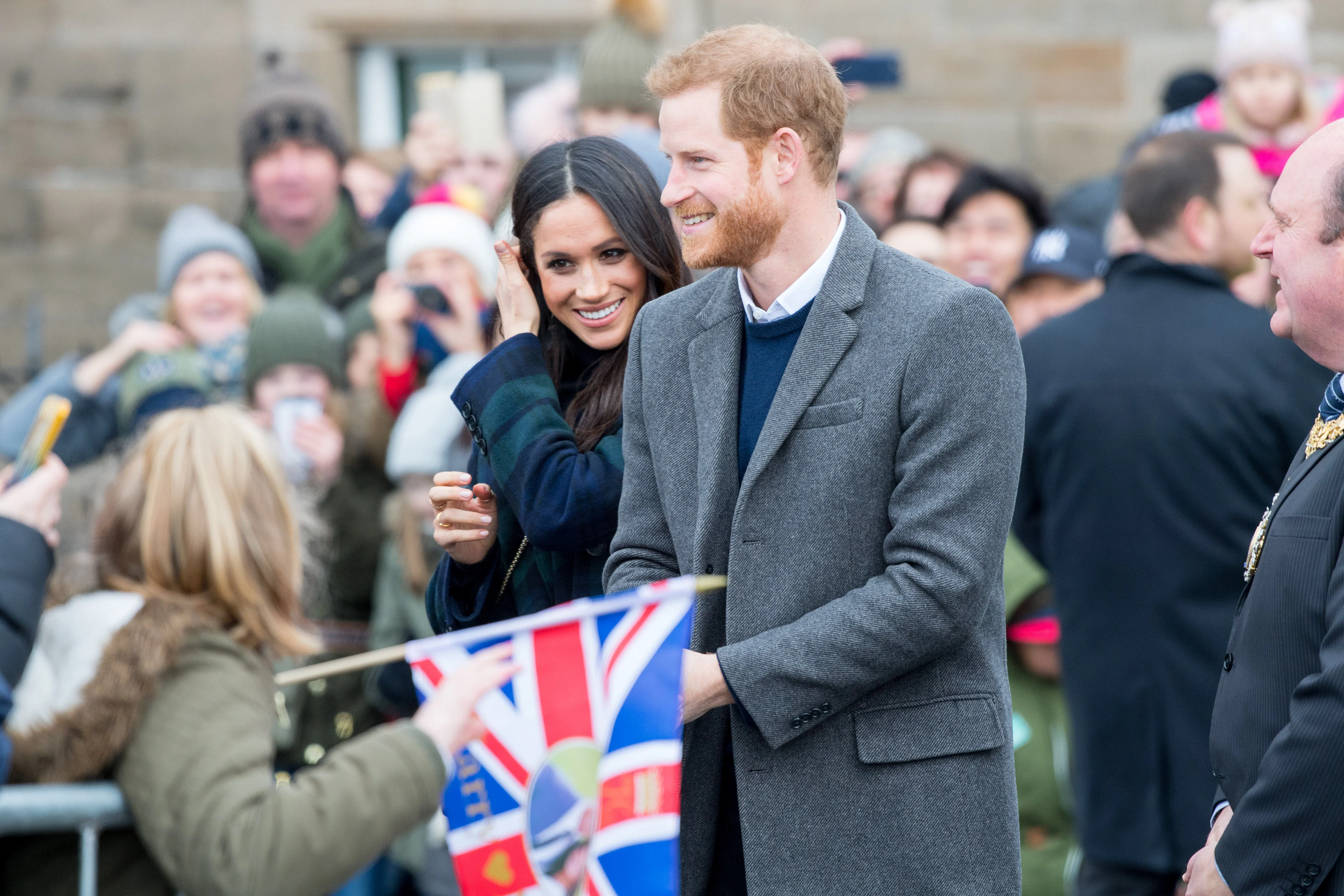 Prince Harry and Meghan Markle interacting with crowds, with Meghan smiling and adjusting her hair, and Harry smiling and looking towards the crowd.