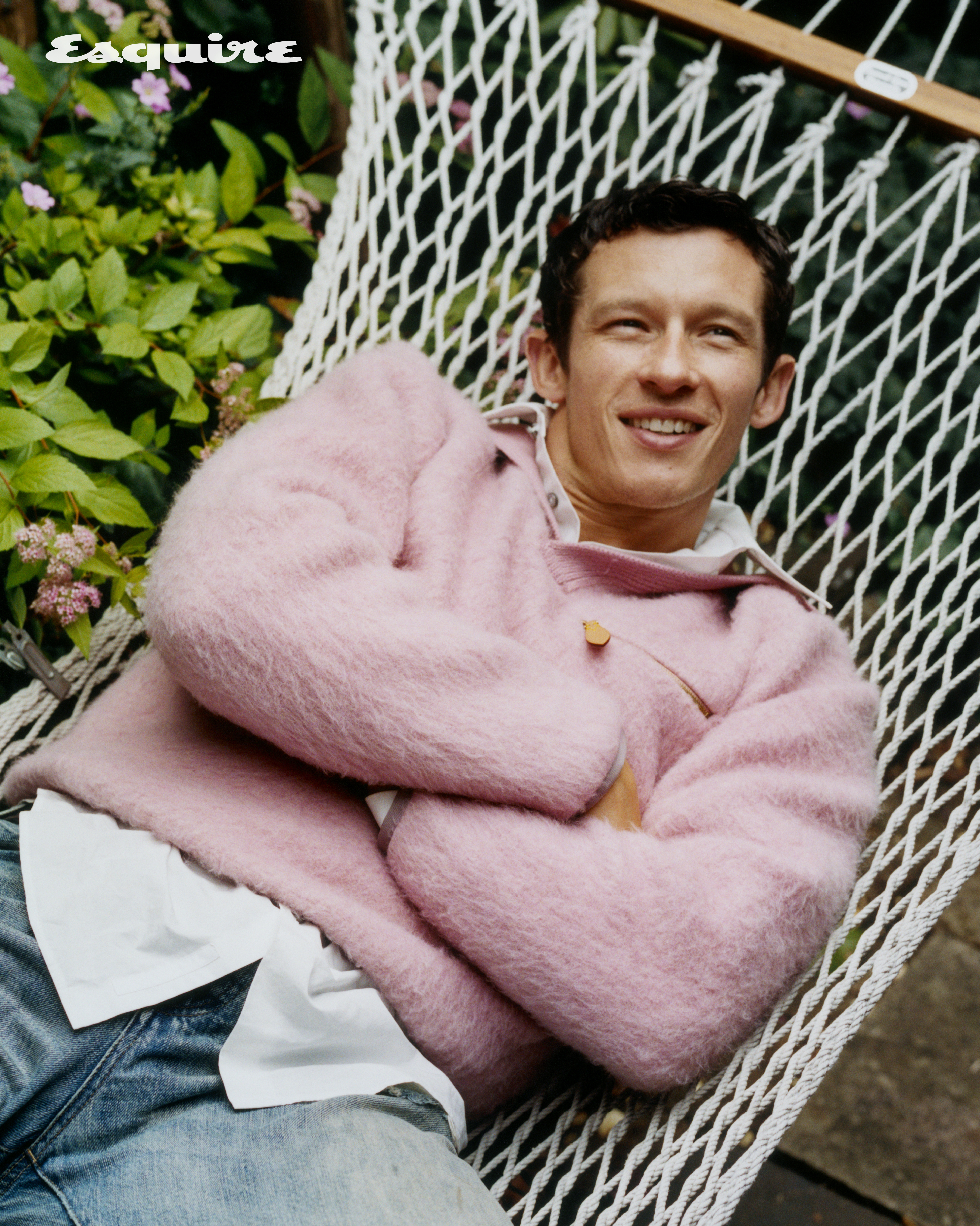 Callum Turner smiling while relaxing in a hammock, wearing a pink fuzzy sweater and blue jeans, with green foliage and pink flowers in the background.