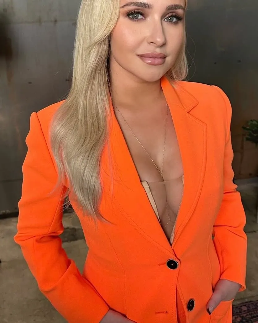 Hayden Panettiere wearing an open orange blazer over a sheer top and a long gold necklace.