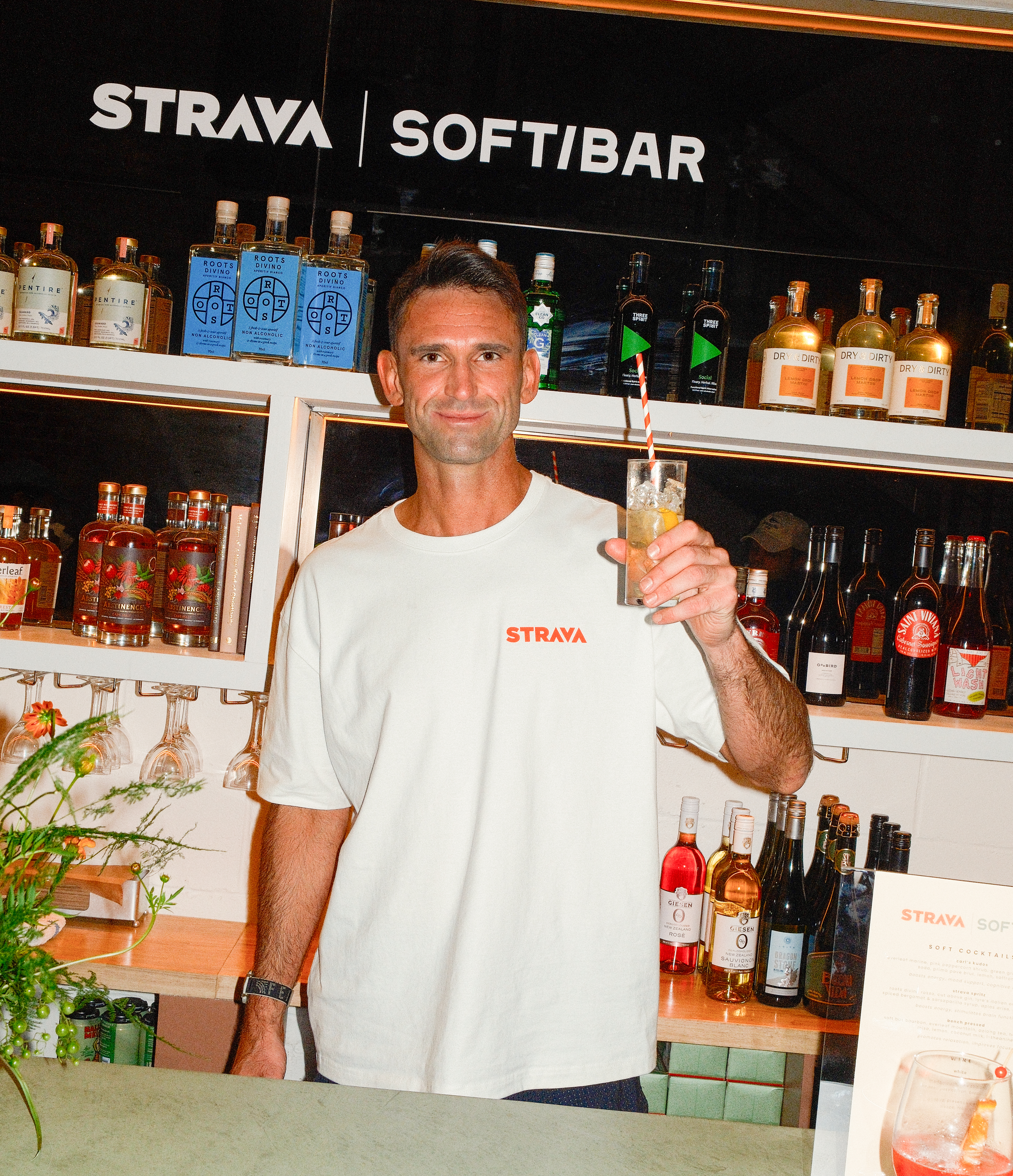 Man in a white "STRAVA" t-shirt holding a drink in front of a bar with "STRAVA SOFTIBAR" written above.