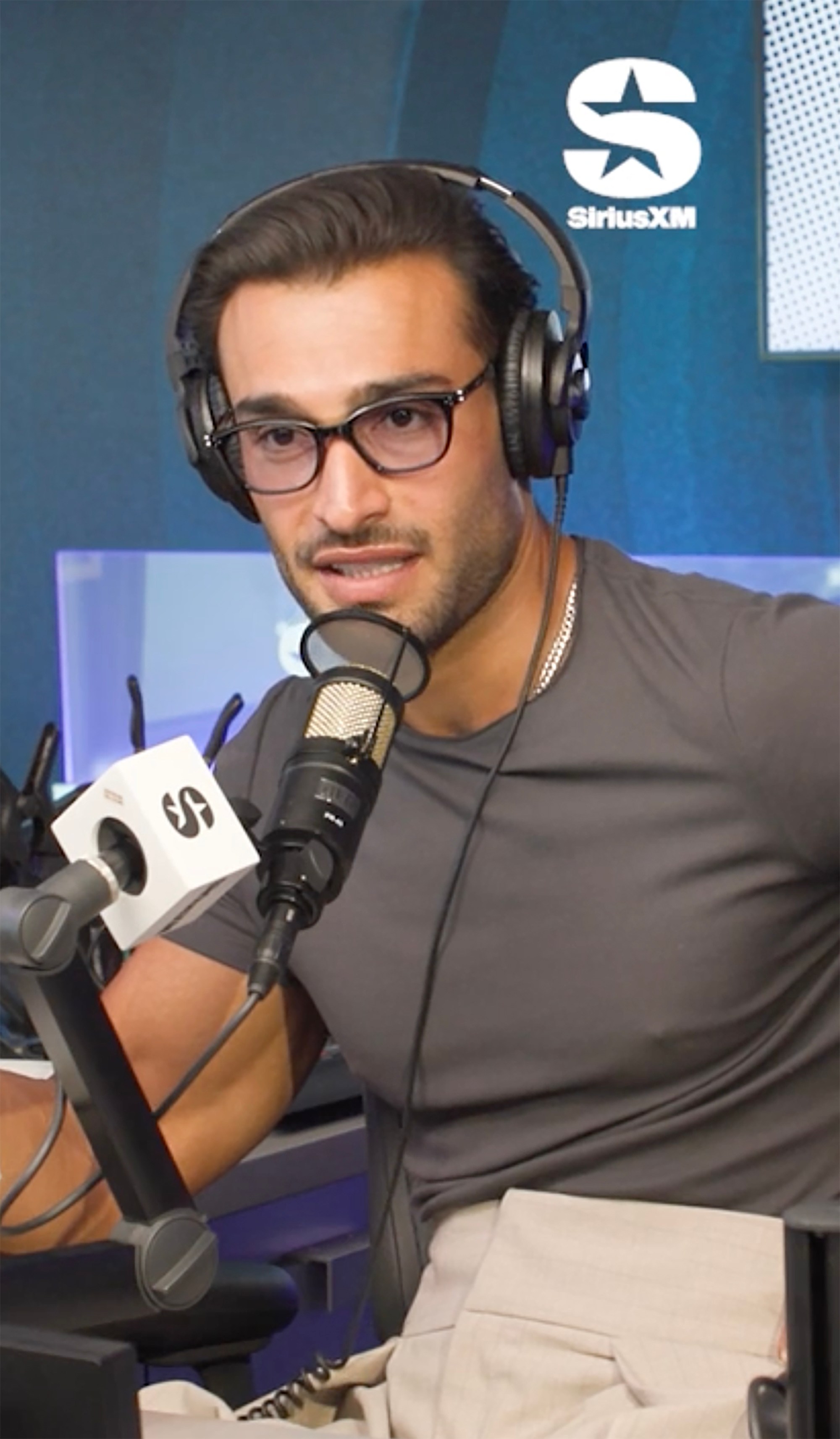 Sam Asghari wearing headphones and glasses, speaking into a microphone with a SiriusXM logo visible.
