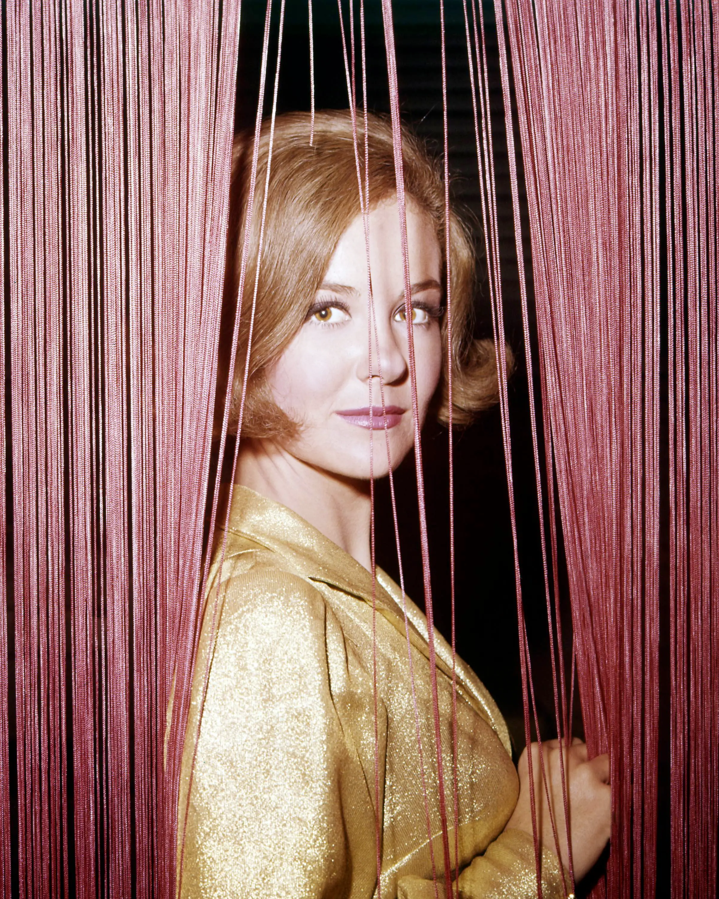 Shelley Fabares in a gold blouse, peering through a red string curtain.