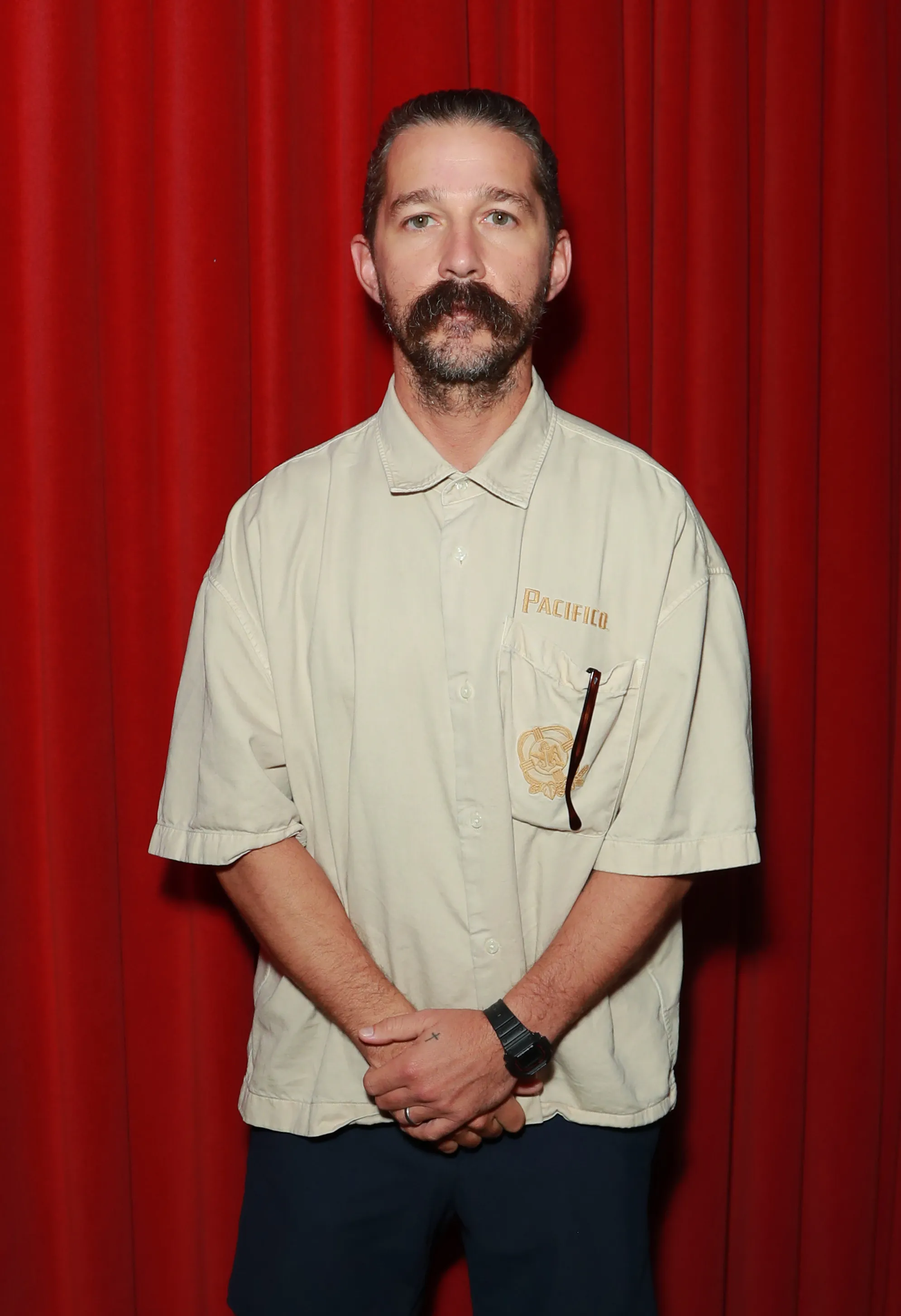 Shia LaBeouf with a mustache and beard, wearing a cream short-sleeved collared shirt and dark pants, standing against a red curtain backdrop.