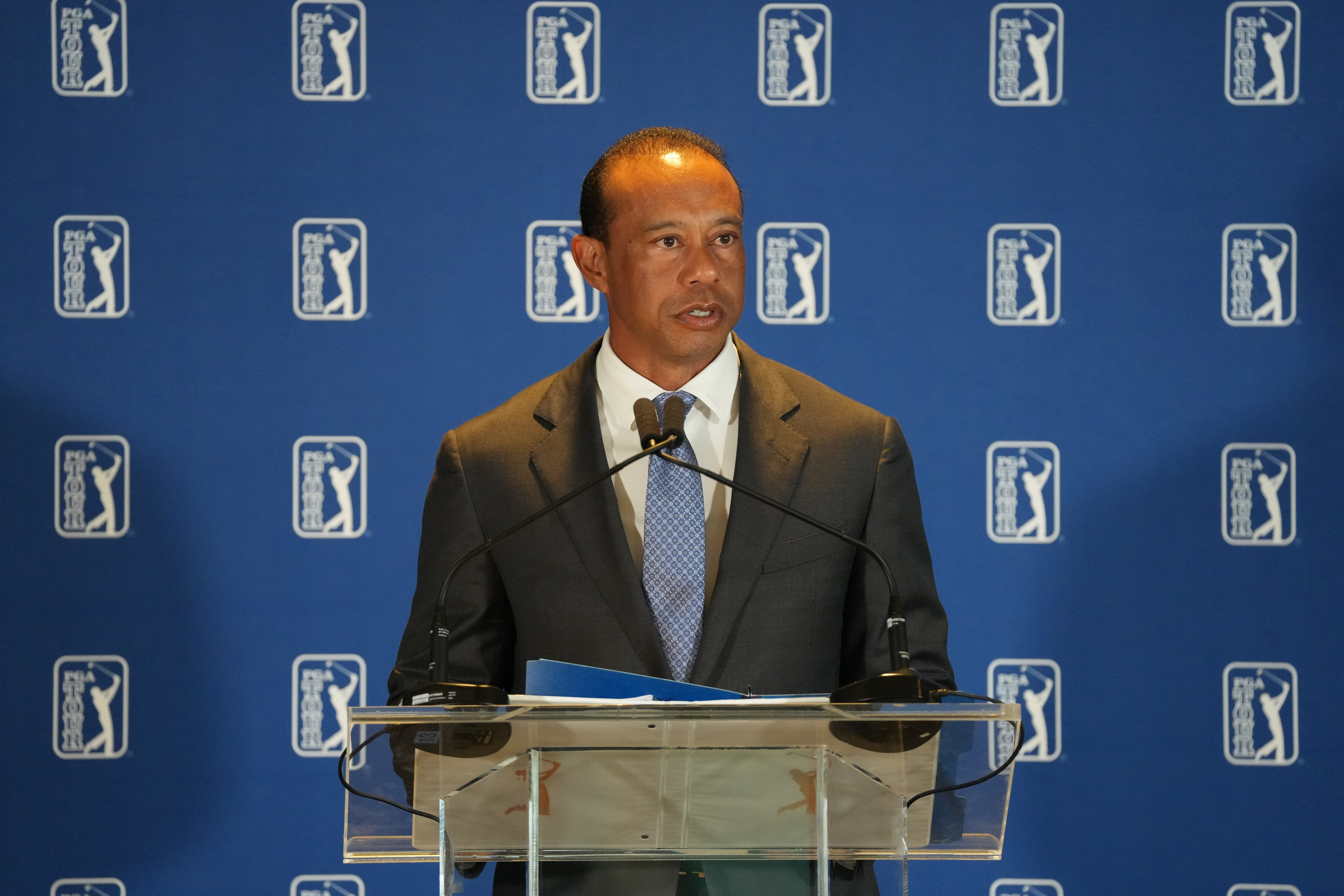 Tiger Woods speaking at a podium with a PGA TOUR backdrop.