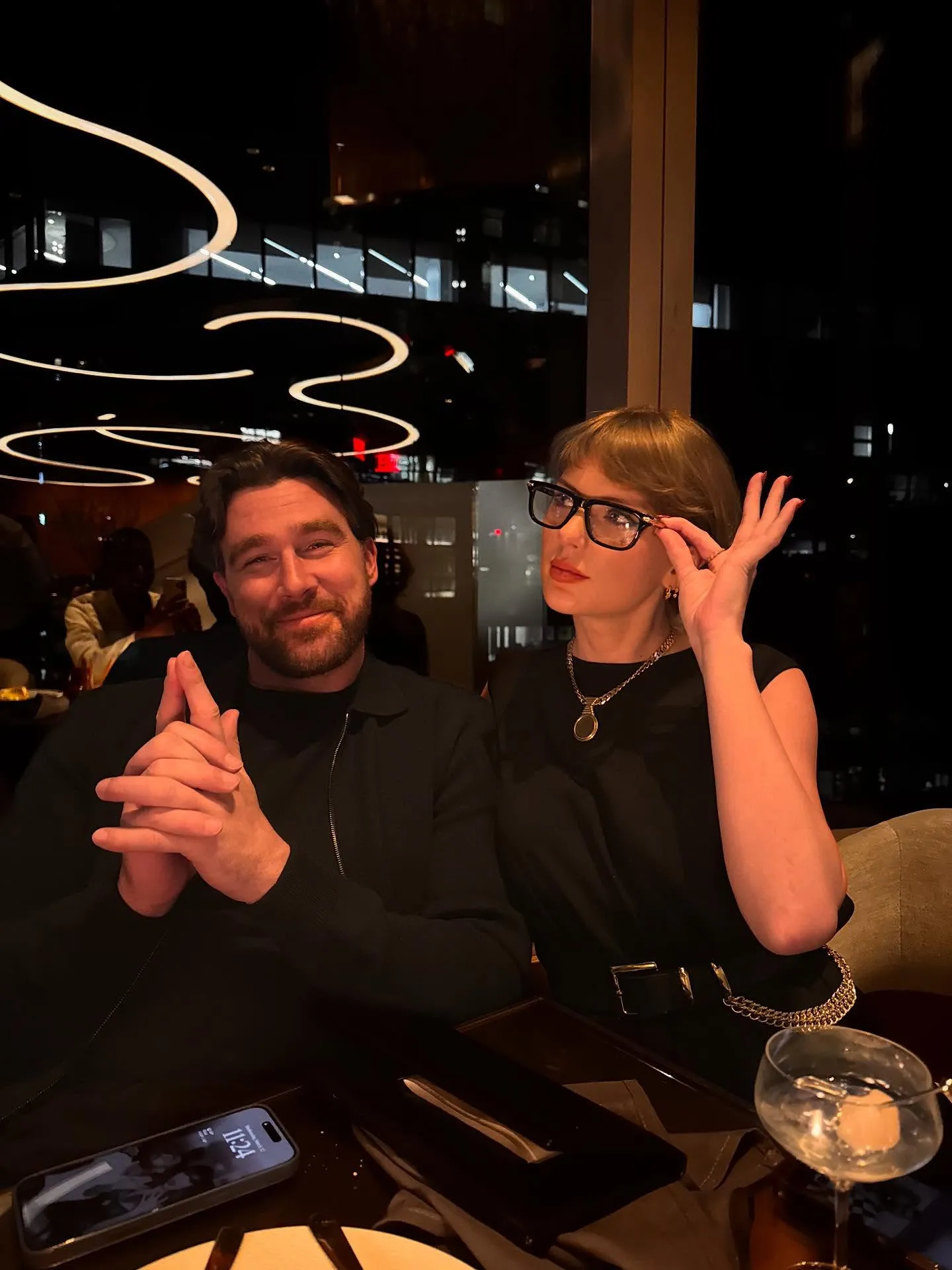 Travis Kelce smiling while sitting next to Taylor Swift, who is adjusting her glasses.