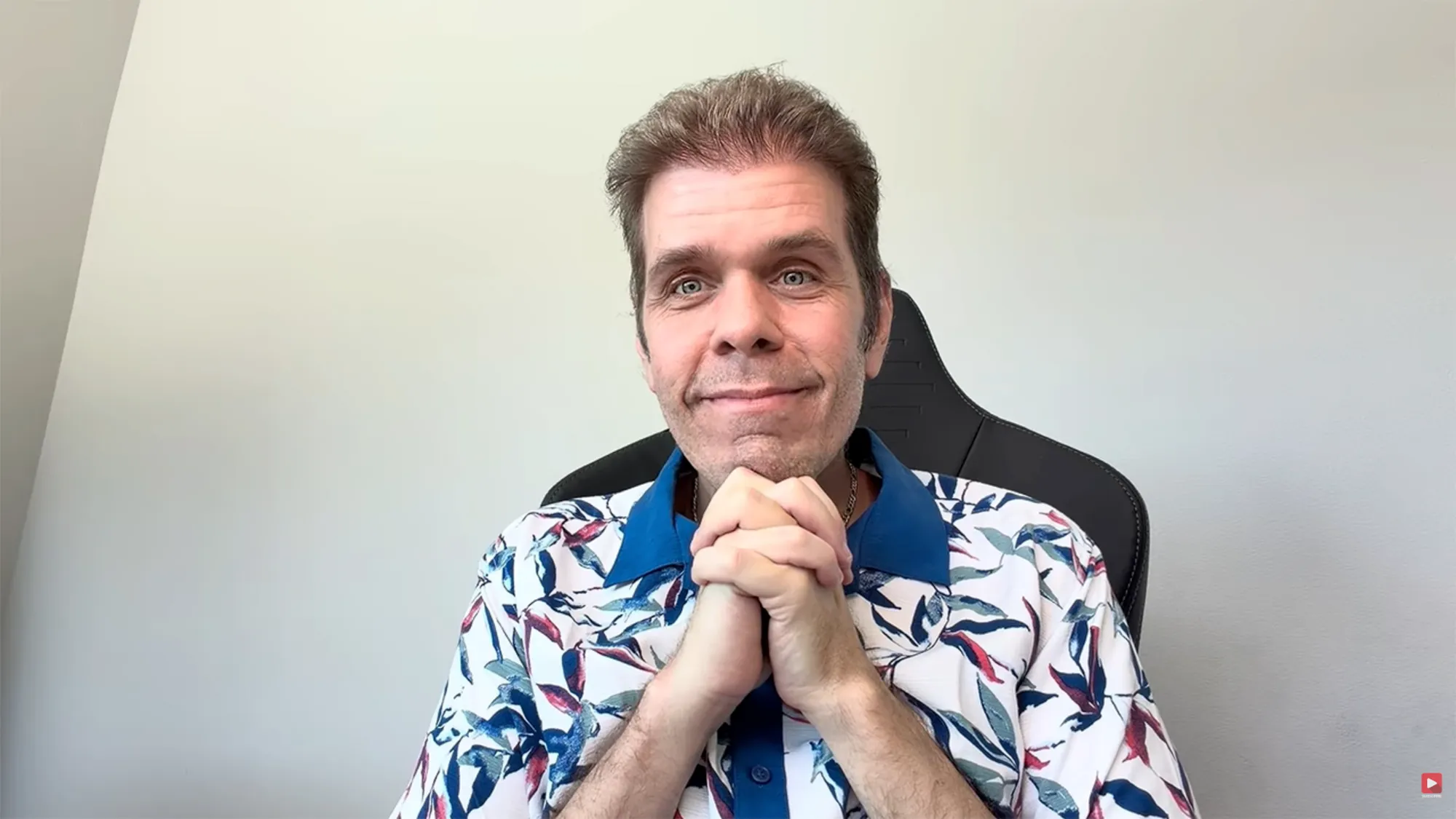 wearing a floral collared shirt, clasping his hands together and looking at the camera.