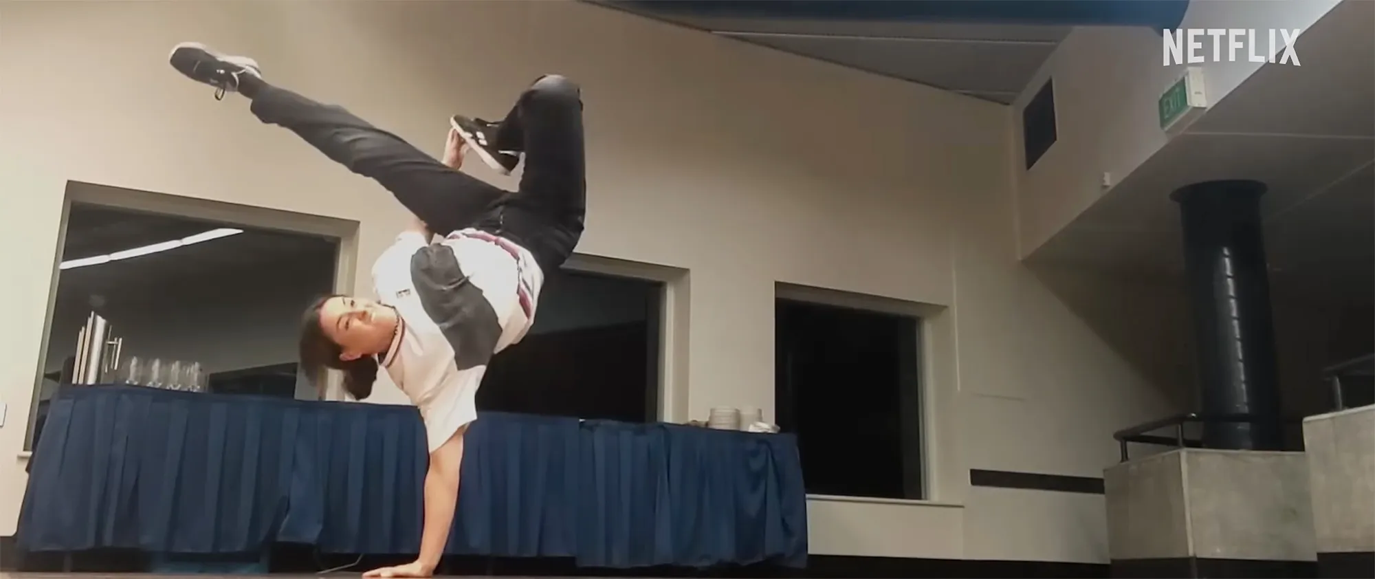 A young woman performing a one-handed handstand with her legs bent in an L-shape.