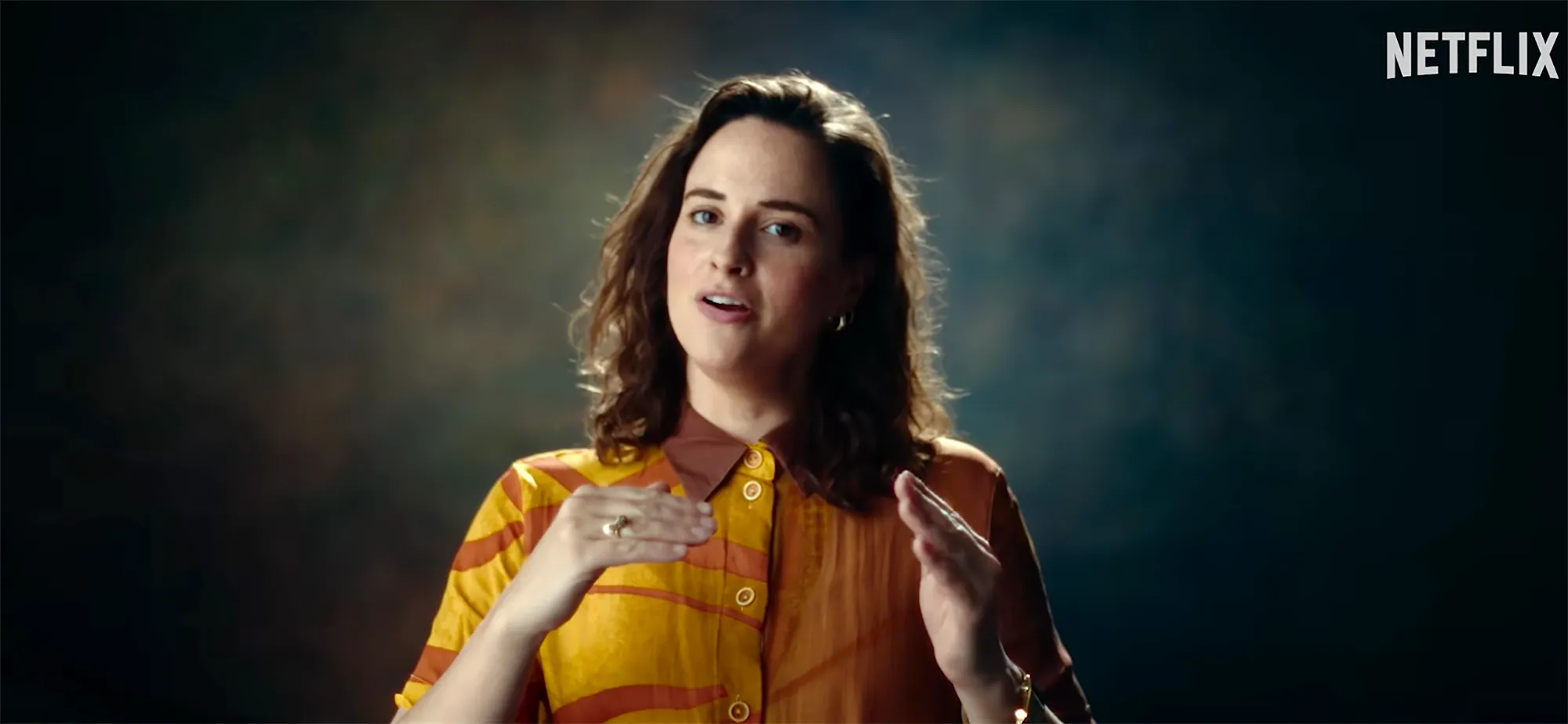 A woman with dark hair wearing a patterned shirt speaks and gestures with her hands.