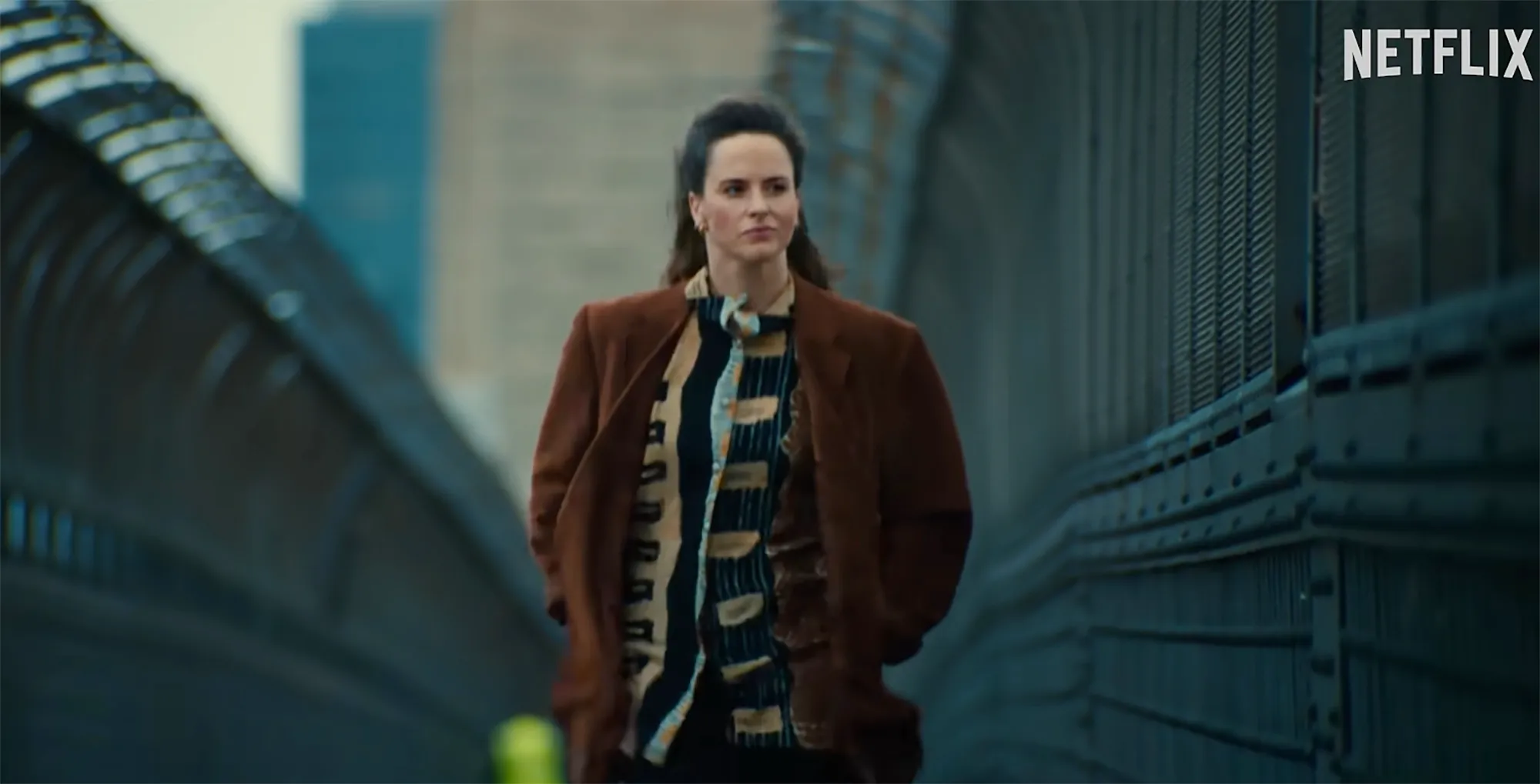 A woman with dark hair in a brown jacket walks on a bridge, with the Netflix logo in the top right.