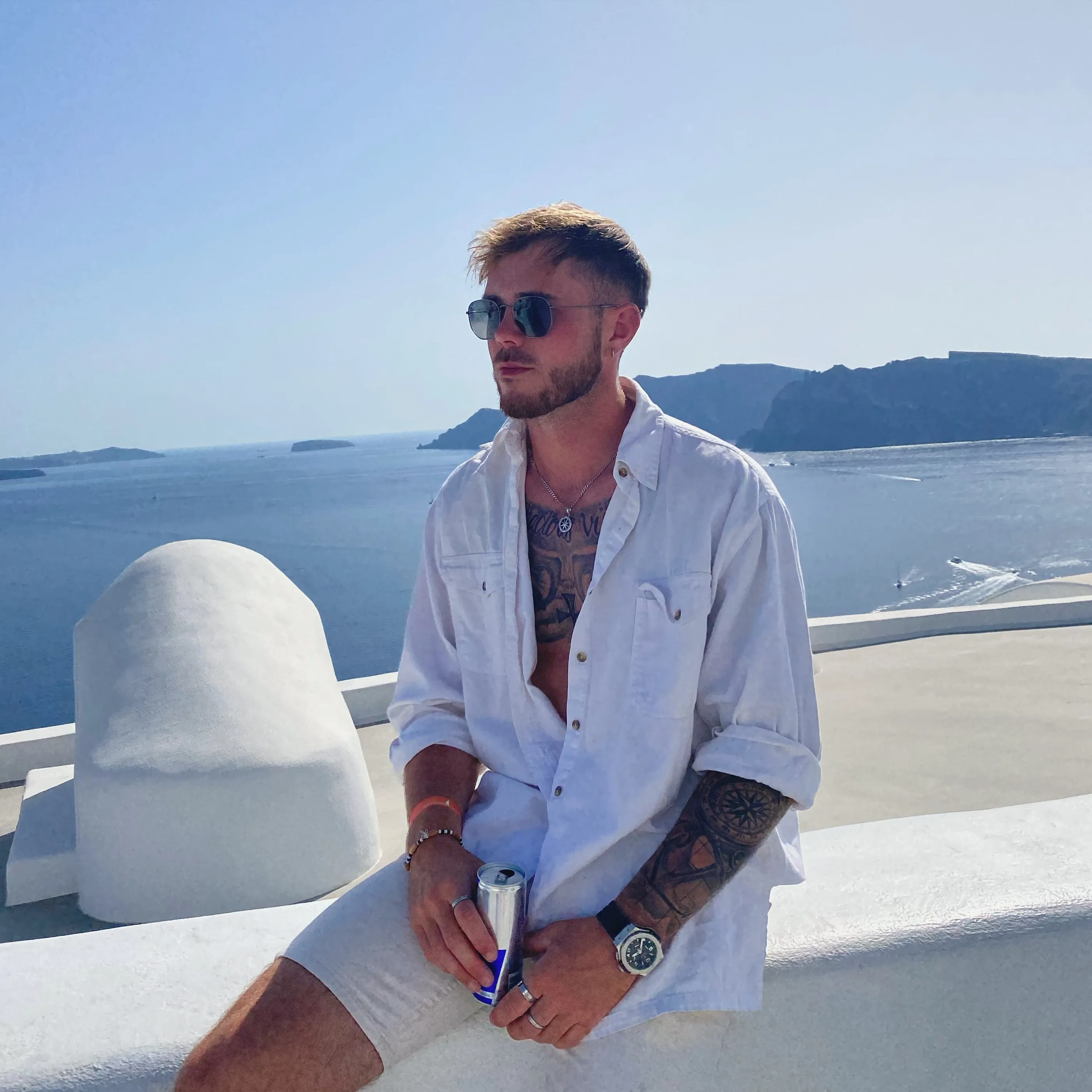 A man in a white shirt and shorts holding a Red Bull can, sitting on a white wall overlooking the ocean with mountains in the background.