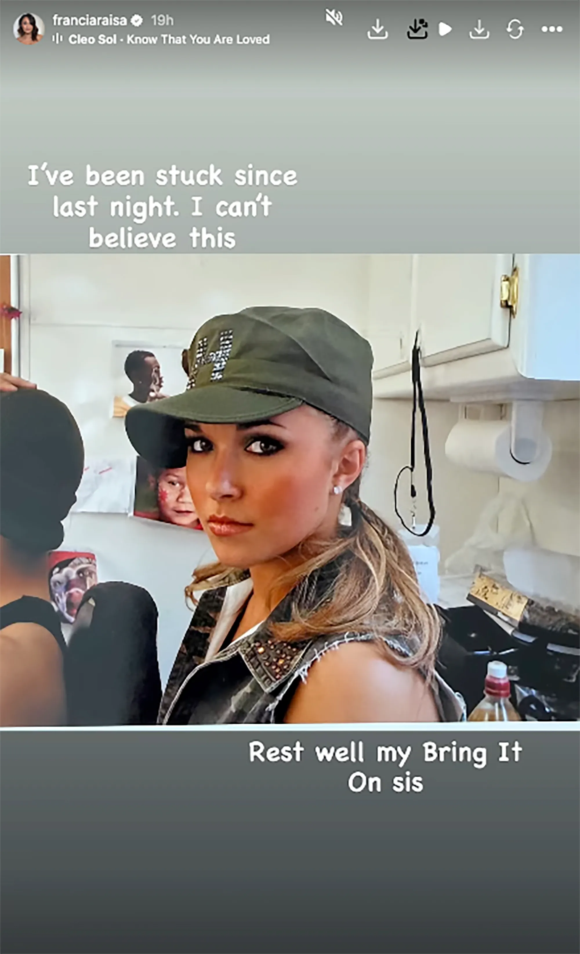 A young woman with a long ponytail, wearing a green cap and a studded vest, looks towards the viewer.
