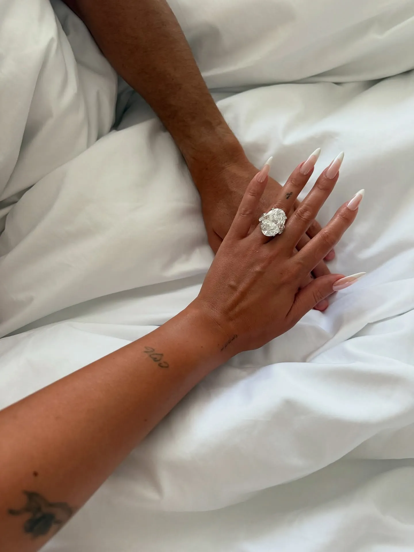 A hand with a large oval engagement ring on the ring finger, holding another hand, both resting on white sheets.