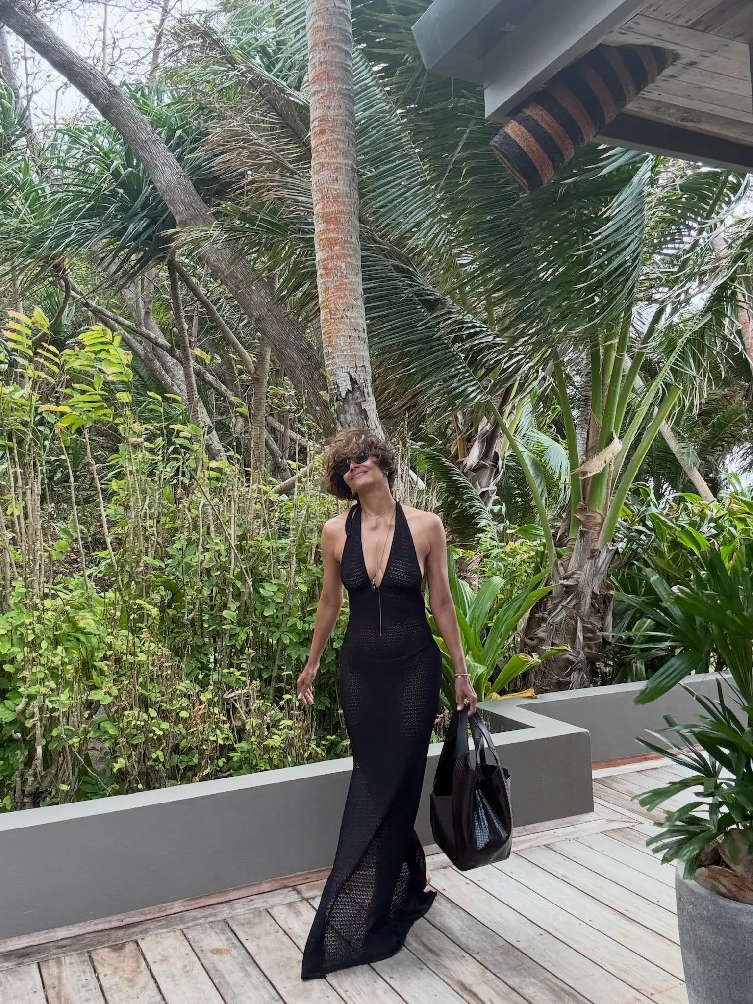 Halle Berry in a black dress, holding a black bag, on a wooden deck surrounded by tropical plants.