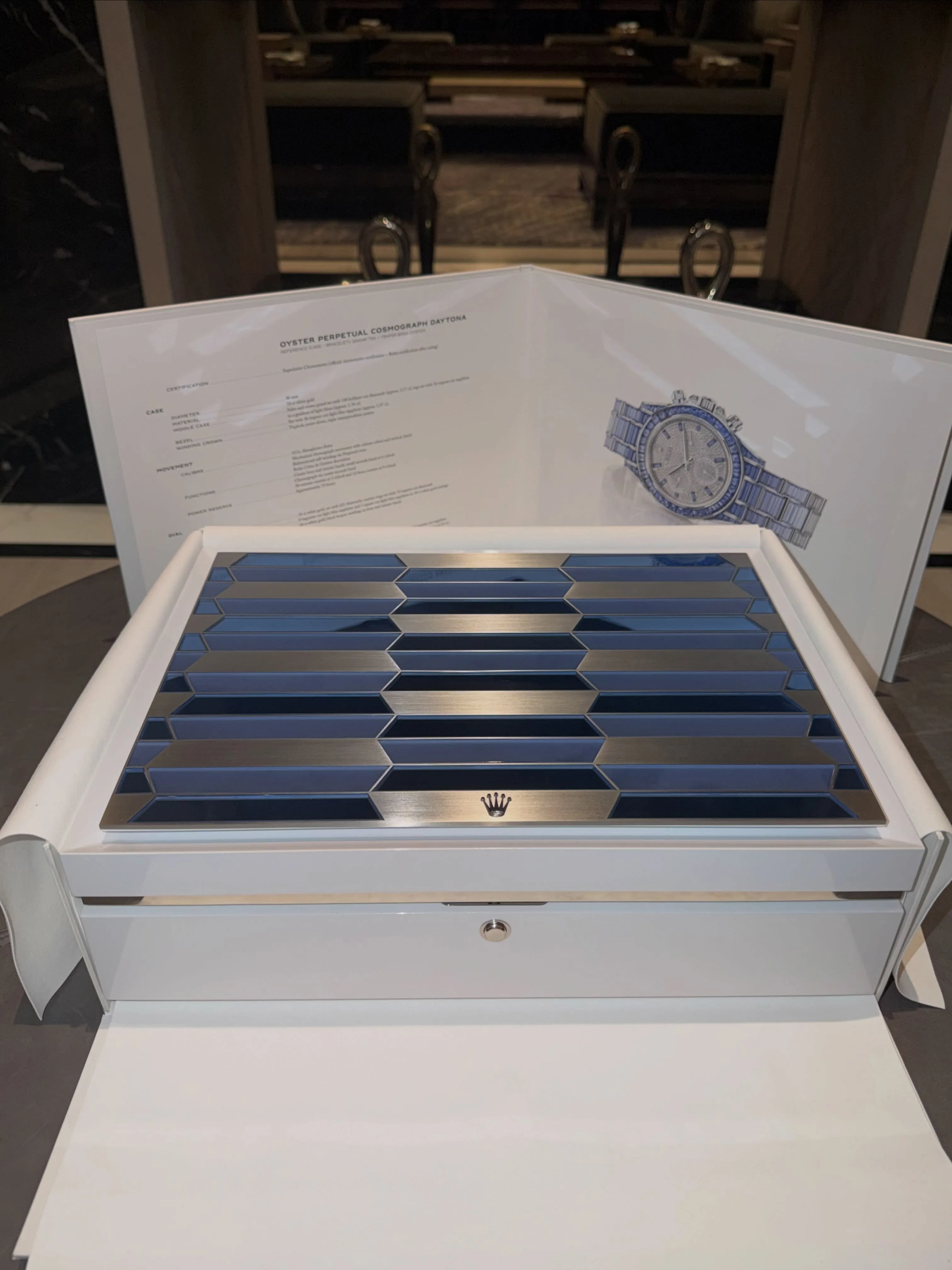 White Rolex box with a blue and silver patterned lid, and an open manual showing an illustration of a diamond-encrusted Rolex Oyster Perpetual Cosmograph Daytona watch.