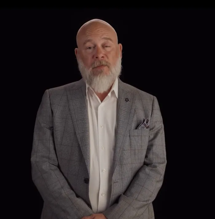 A bald man with a white beard, wearing a gray plaid suit and white shirt, stands against a black background.