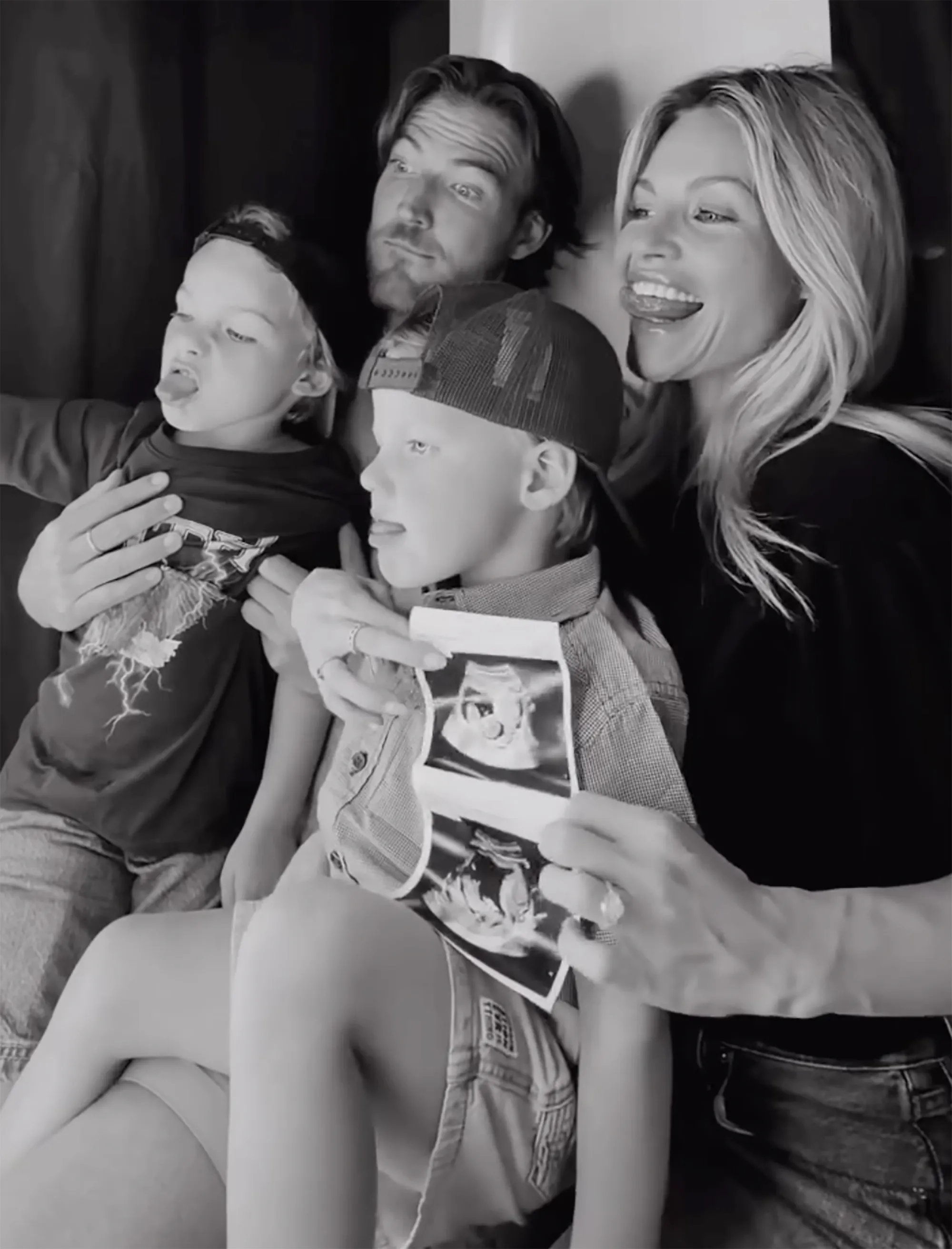 Black and white photo of a family of four, with two young children and their parents, posing for a photo booth picture. The mother is holding up ultrasound images.