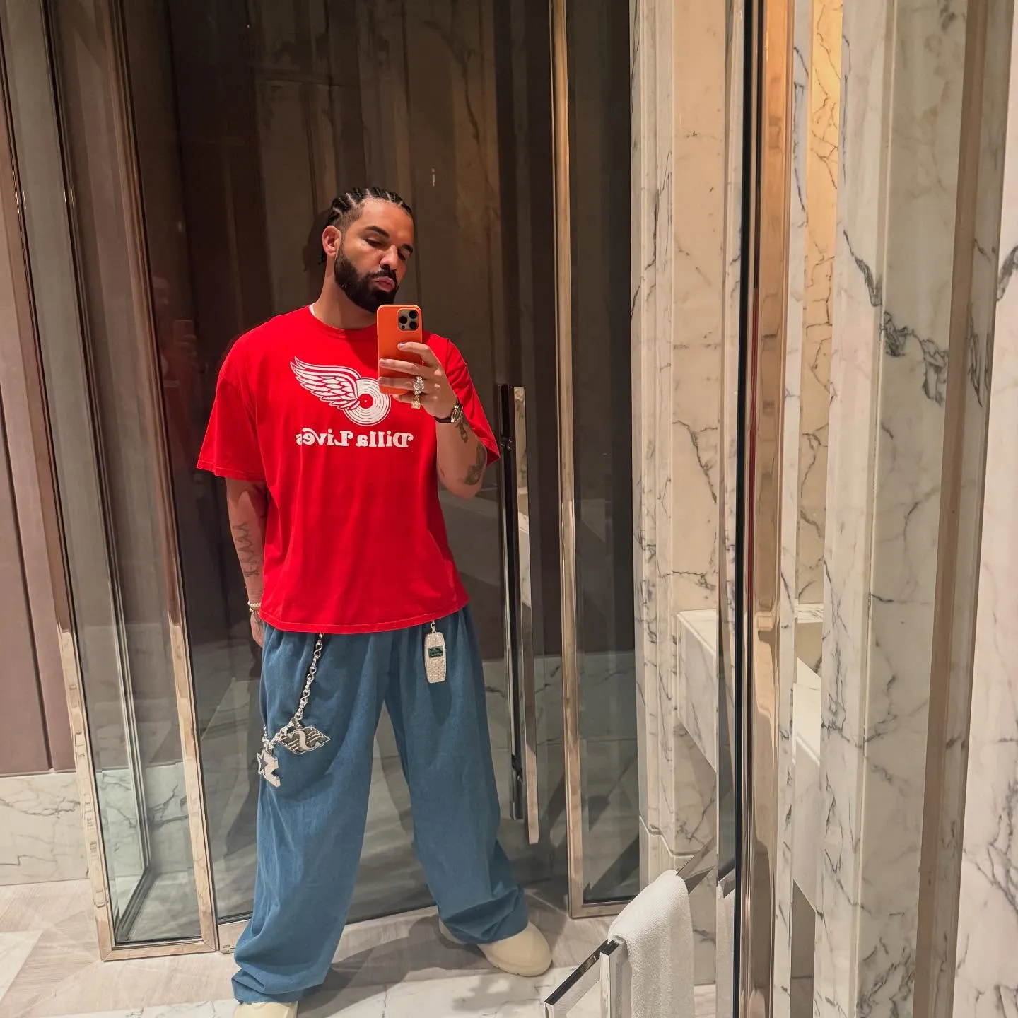 Drake poses for a mirror selfie, wearing a red T-shirt and baggy blue jeans.