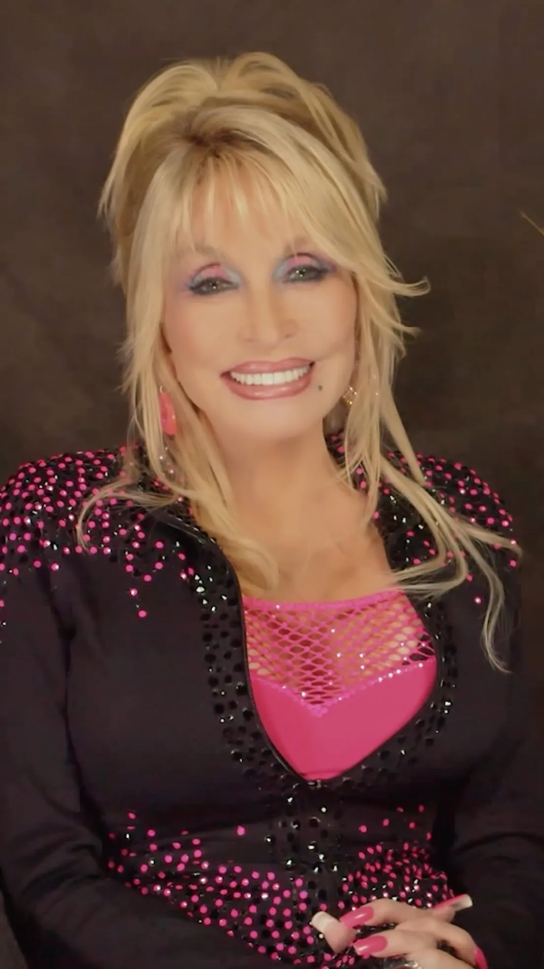Dolly Parton smiling, wearing a black jacket with pink and black sequins and a bright pink mesh top.