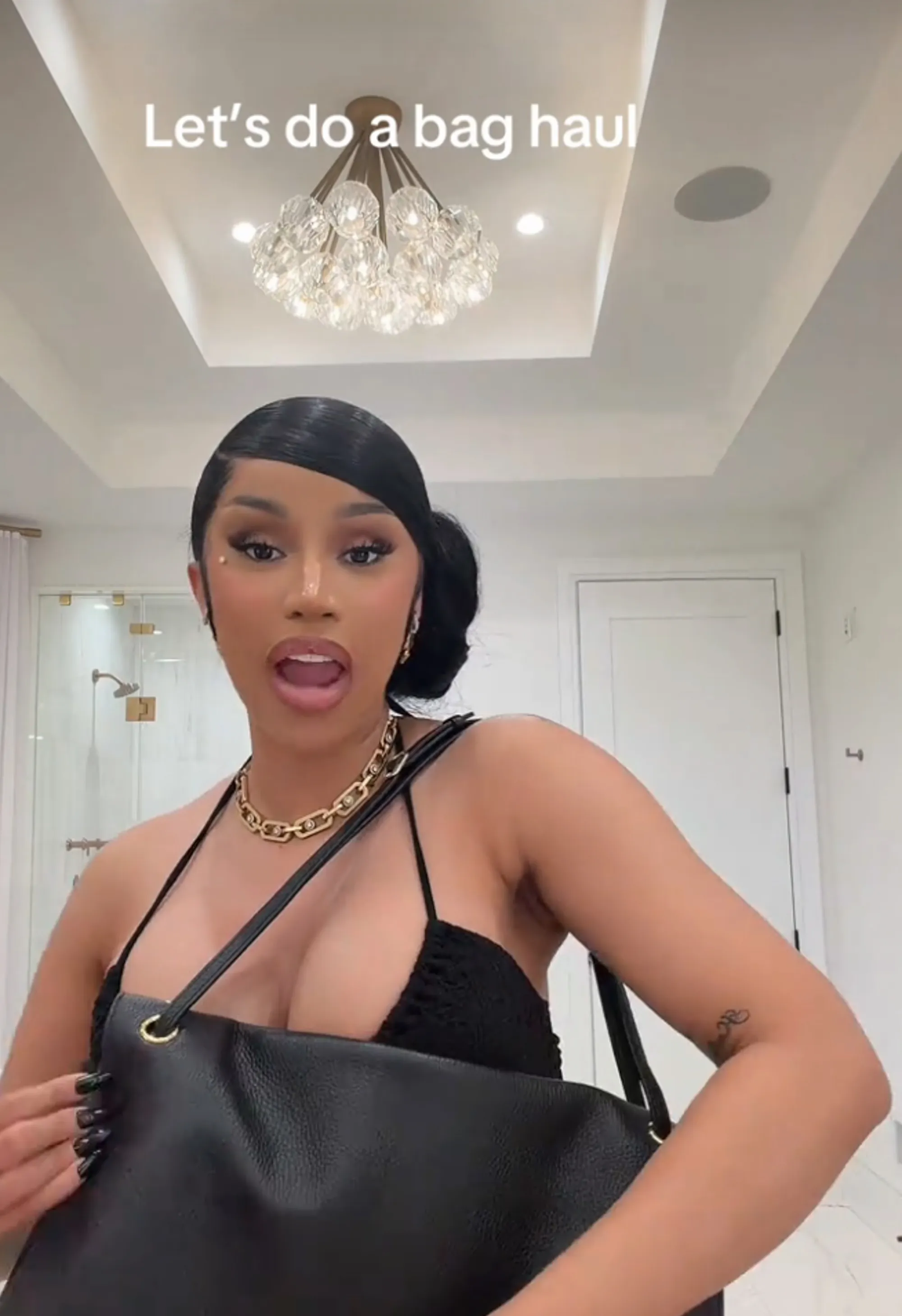 Cardi B in a black top and a gold chain necklace, holding a black handbag.