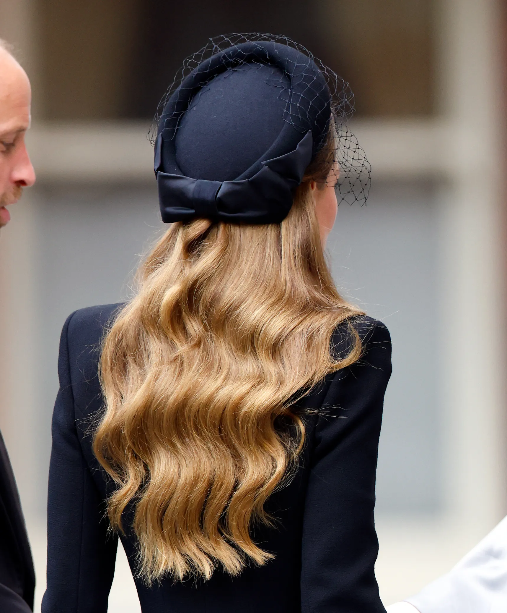 Catherine, Princess of Wales, wearing a black pillbox hat with a bow and net, stands next to an unidentified man.