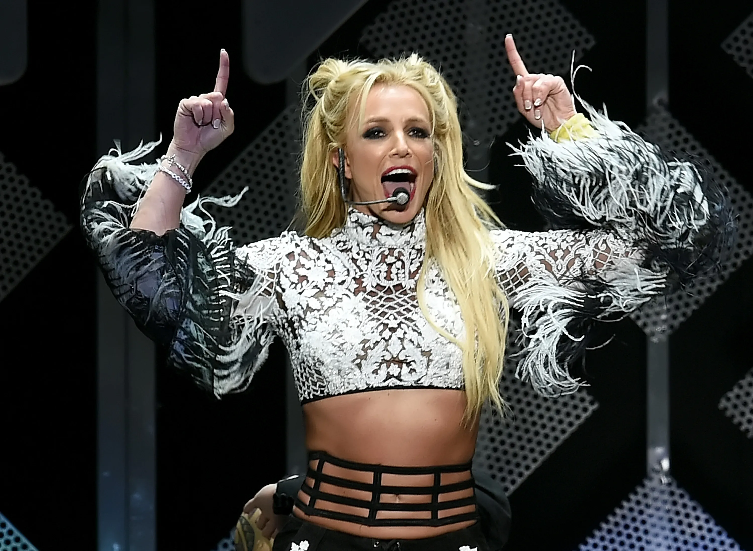Britney Spears performing with her hands raised, wearing a white lace crop top with feathery sleeves.