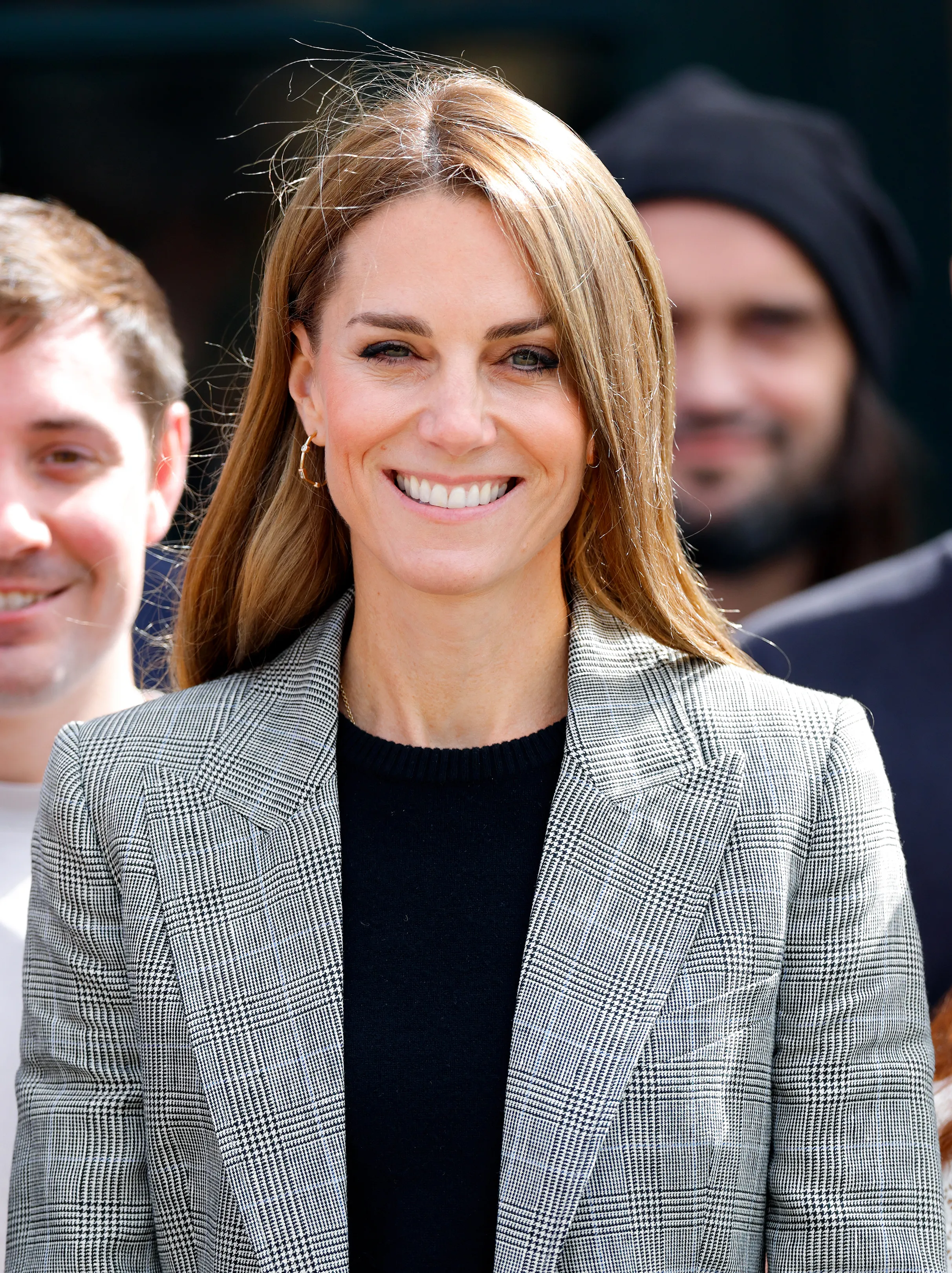 Catherine, Princess of Wales, smiles, wearing a grey plaid jacket over a black top, with gold hoop earrings and blonde hair.