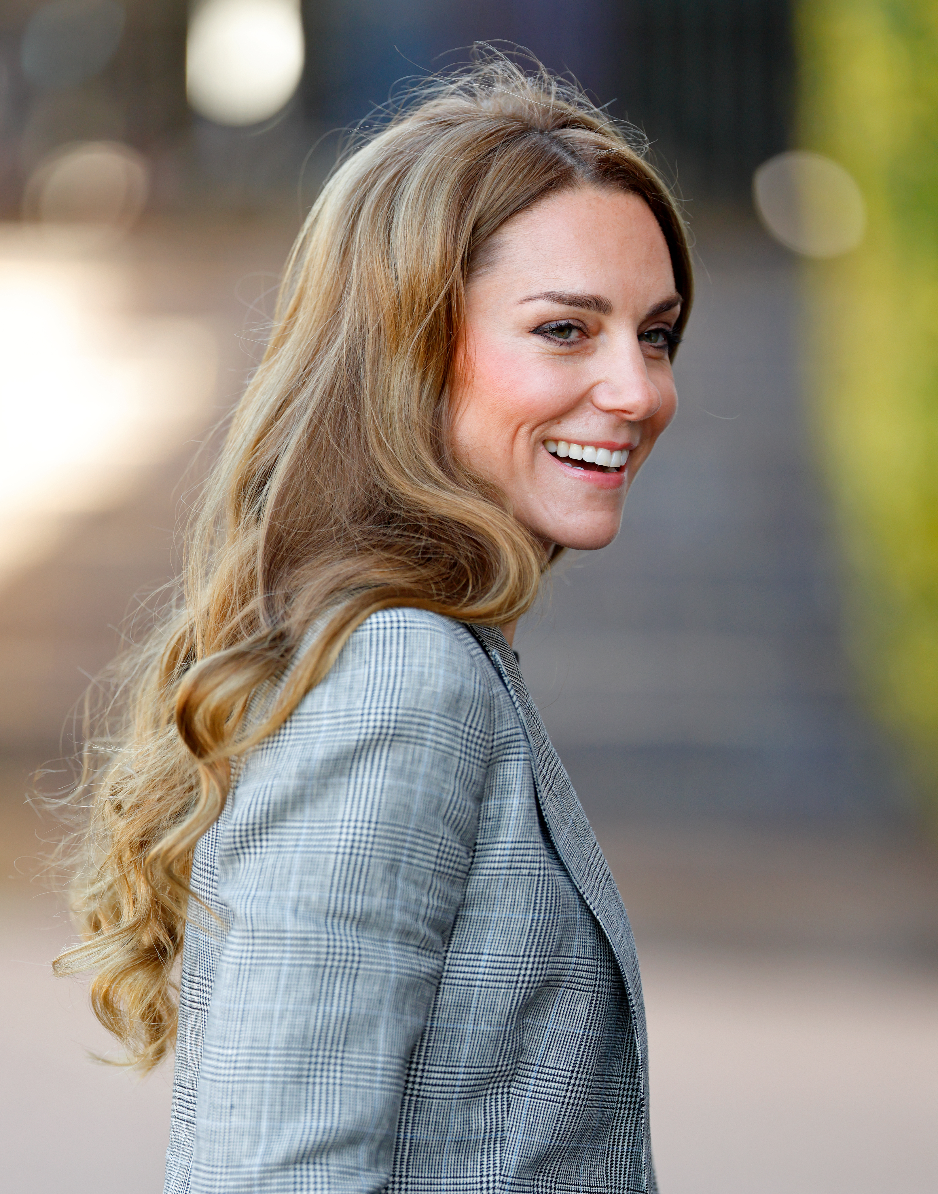 Catherine, Princess of Wales, smiles over her shoulder, wearing a grey plaid jacket.