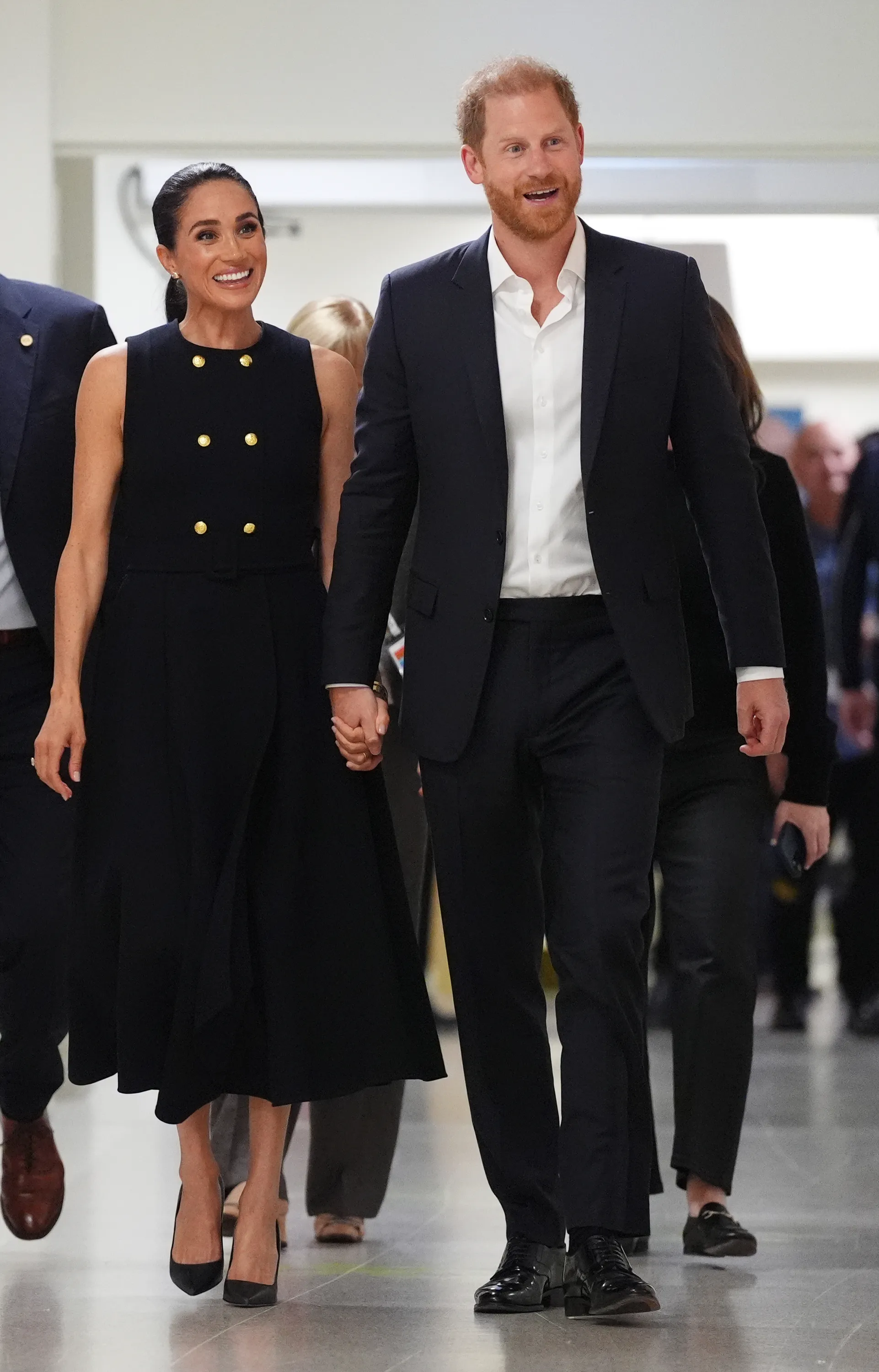 Meghan, Duchess of Sussex, and Prince Harry, Duke of Sussex, holding hands and smiling while walking.
