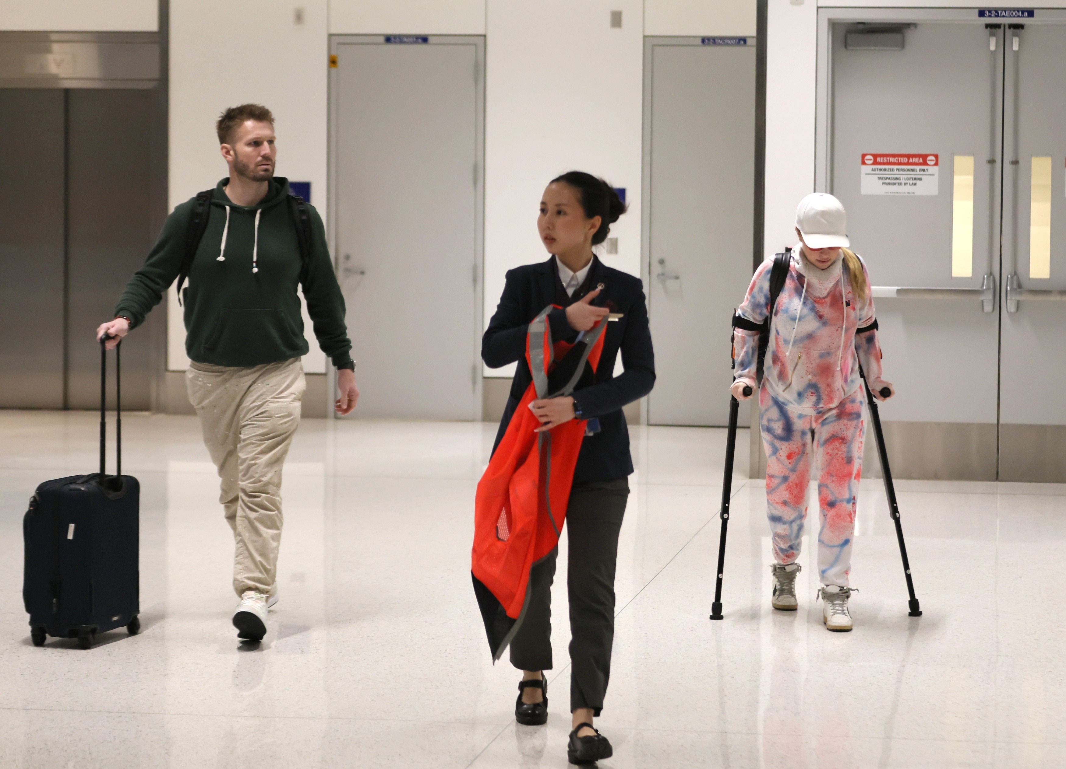 Hayden Panettiere on crutches in a tie-dye tracksuit, walking with Brian Hickerson and an airport employee.