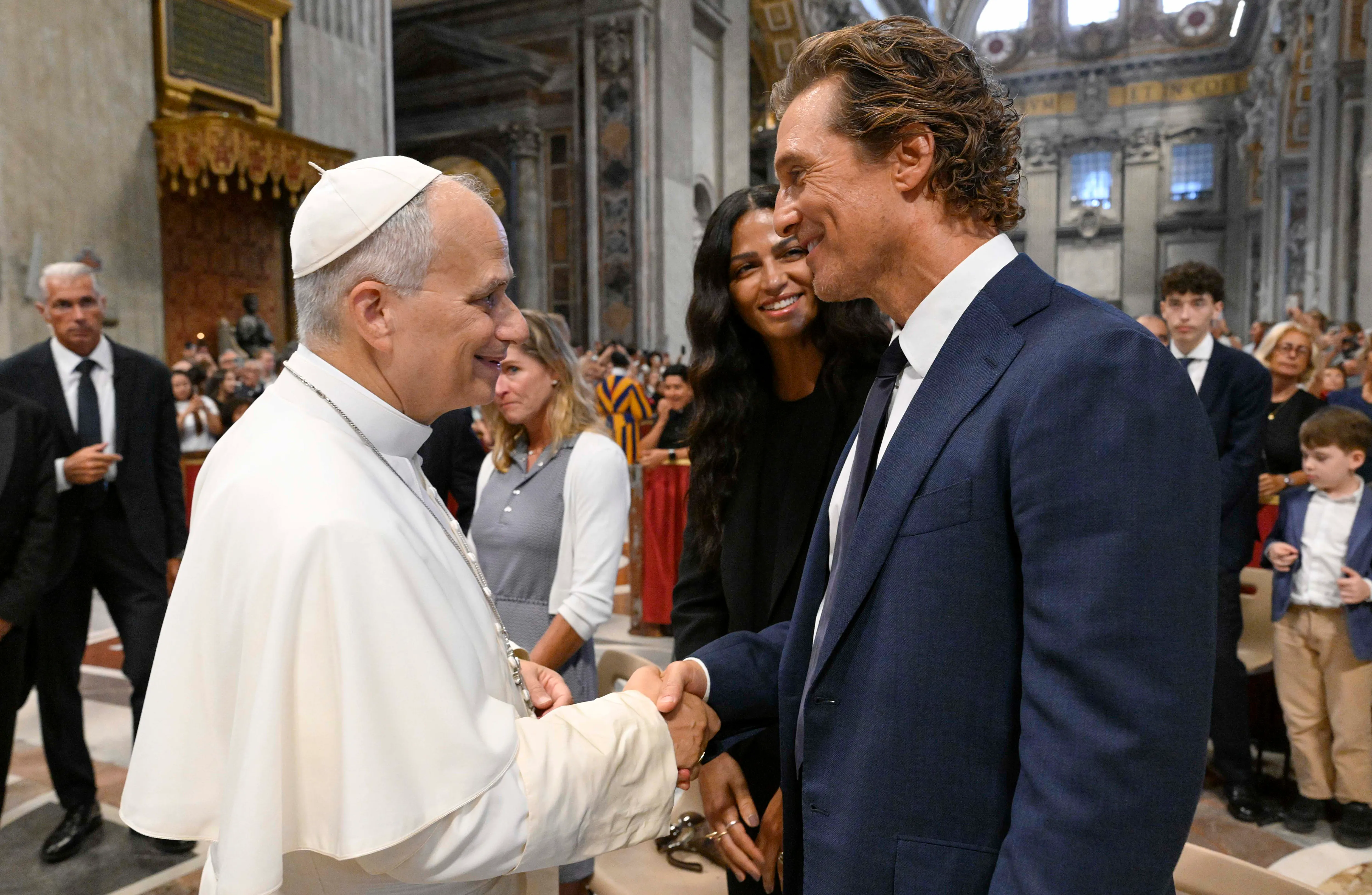Pope Leo XIV shaking hands with Matthew McConaughey, while Camila Alves stands smiling behind McConaughey.