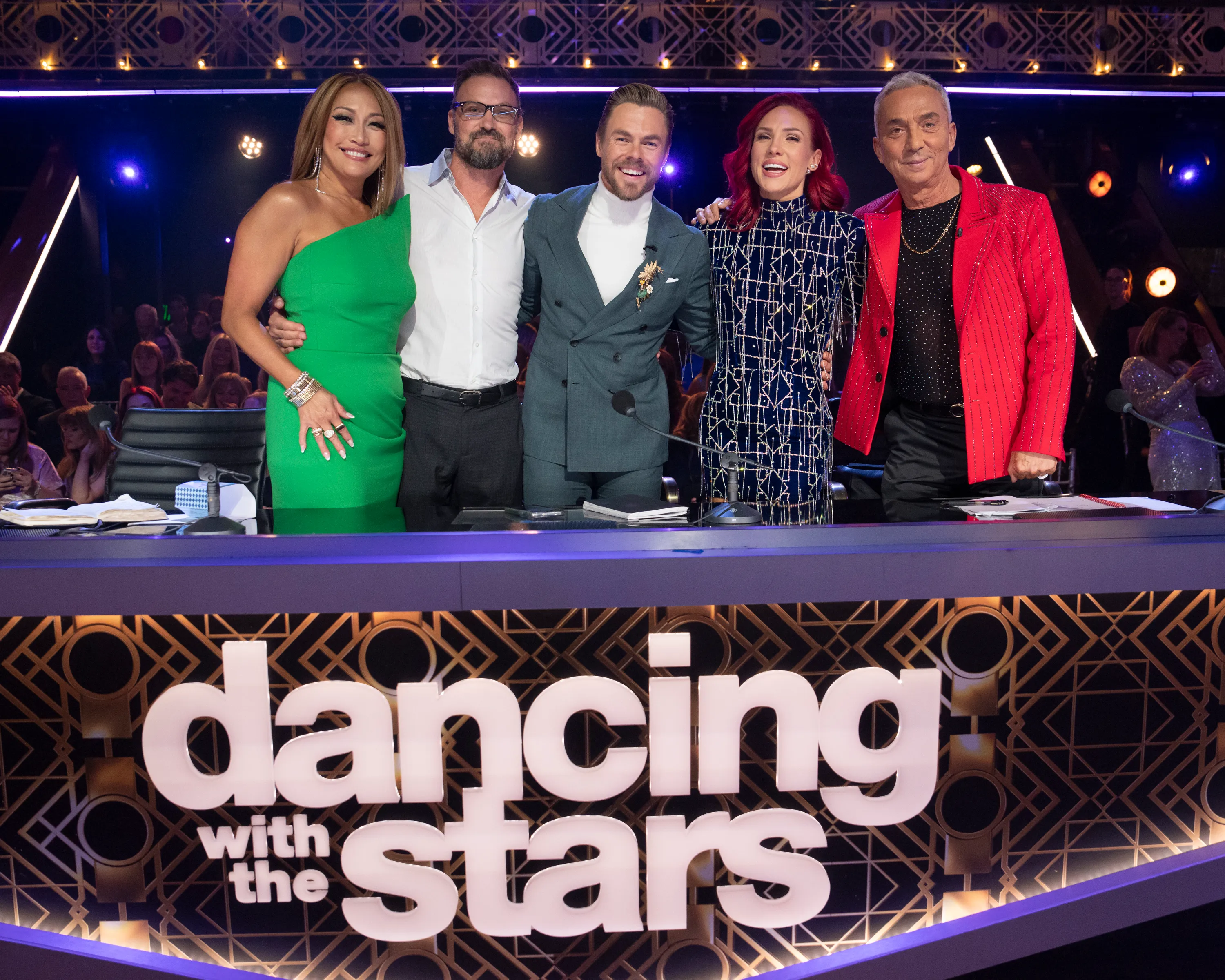 "Dancing with the Stars" judges and hosts Carrie Ann Inaba, Brian Austin Green, Derek Hough, Sharna Burgess, and Bruno Tonioli stand behind a desk with the show
