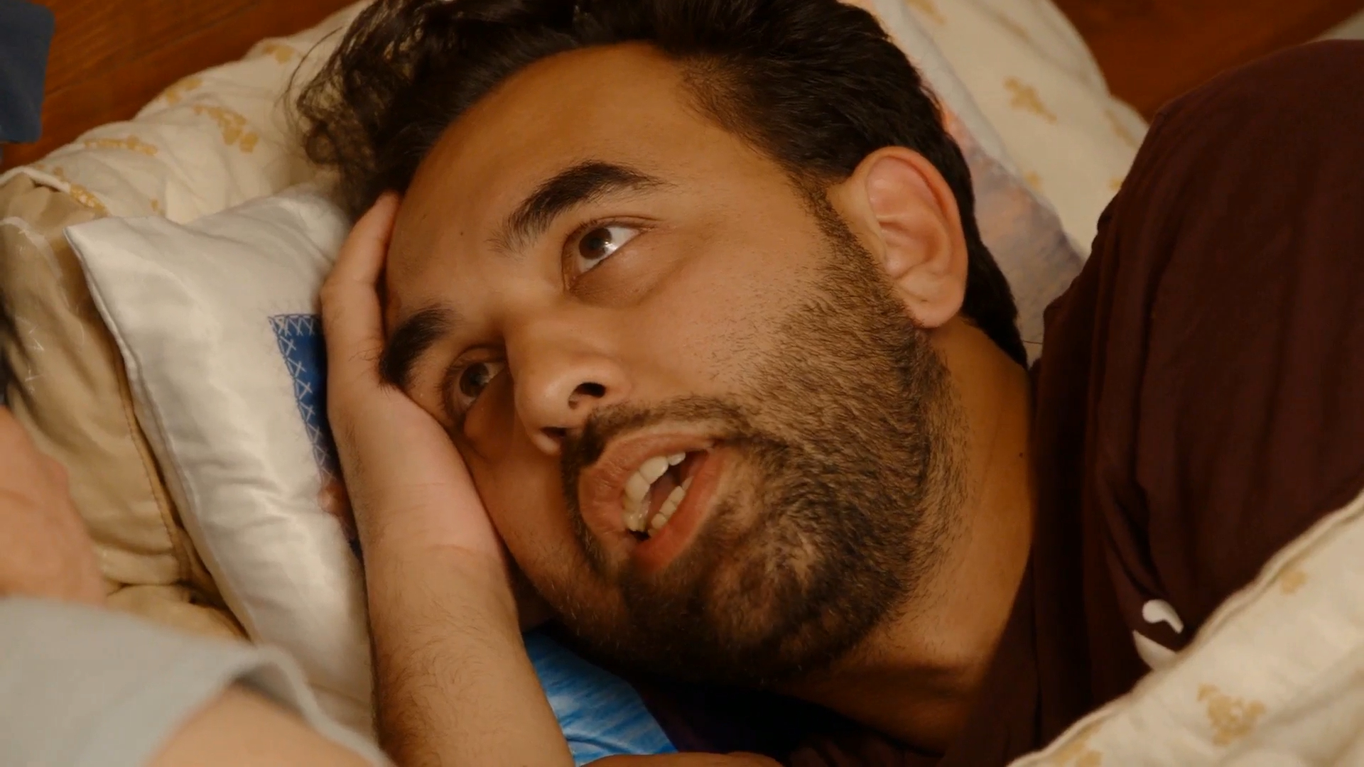 A man with dark hair and a beard, lying in bed with his head on a pillow, looks up and to the left with an open mouth.