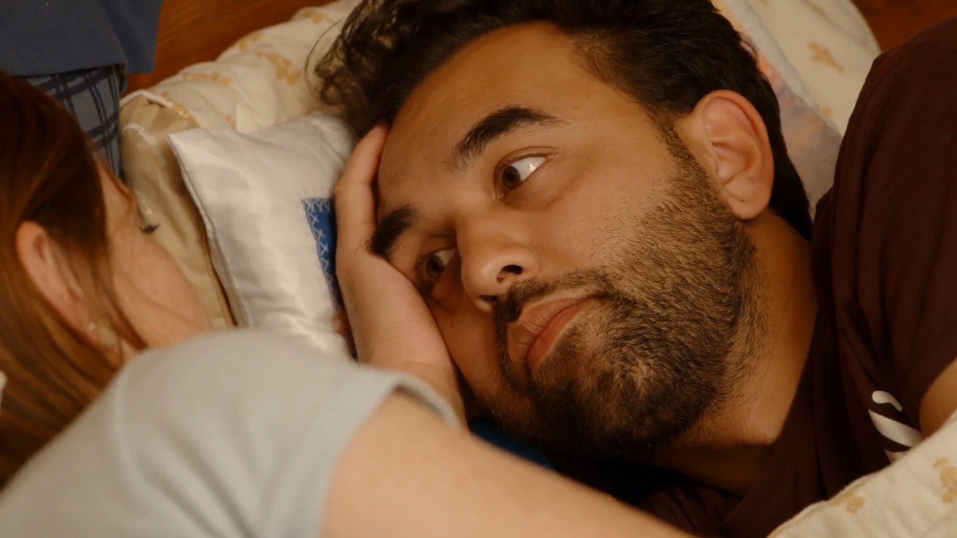 Man with a beard looks surprised while lying in bed next to a woman.