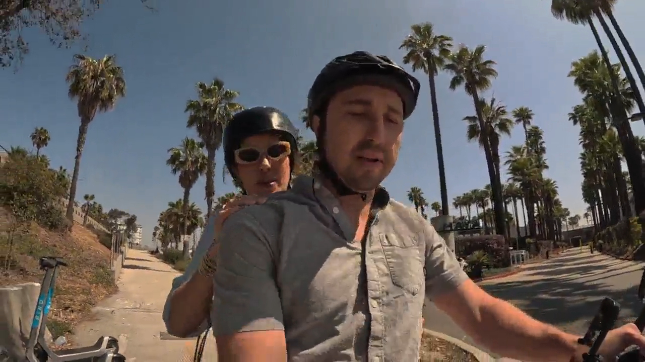 A man and woman wearing helmets riding on a scooter on a sunny day with palm trees in the background.