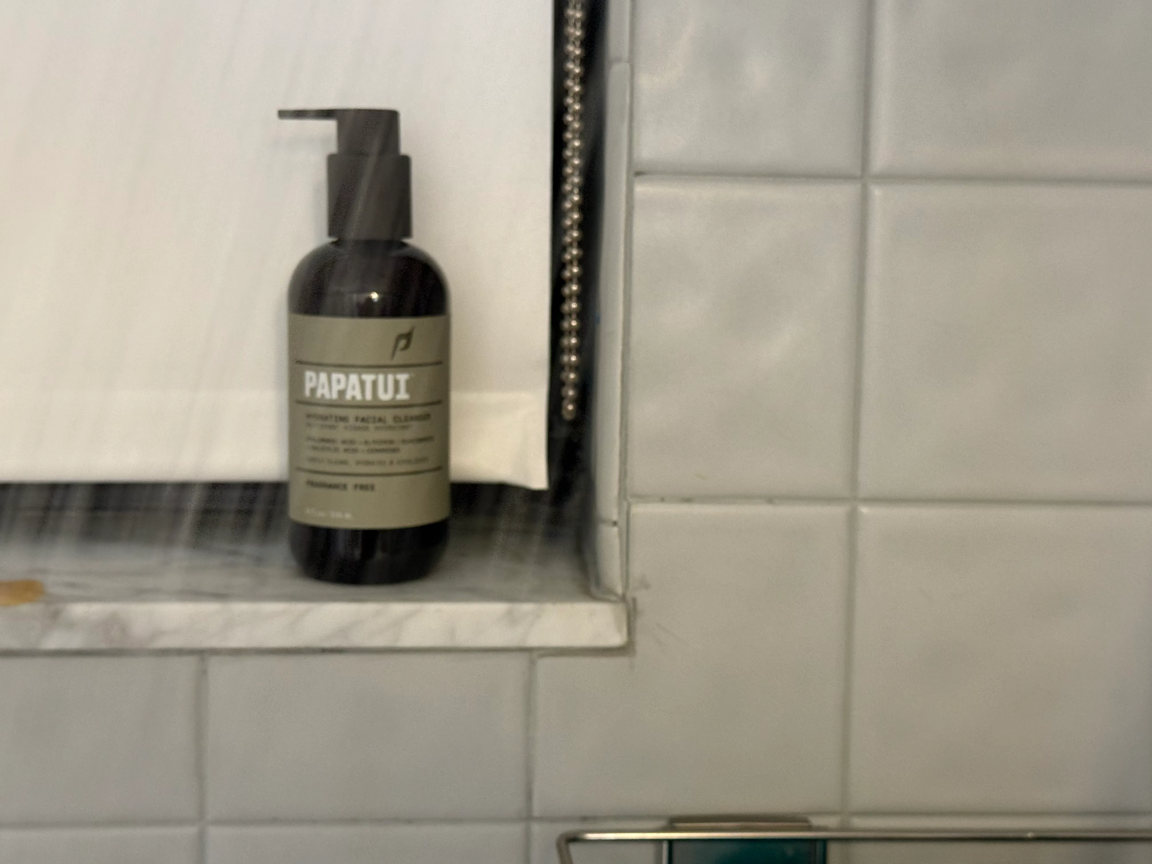 A dark bottle of Papatui Hydrating Facial Cleanser with a pump top sits on a tiled shelf.