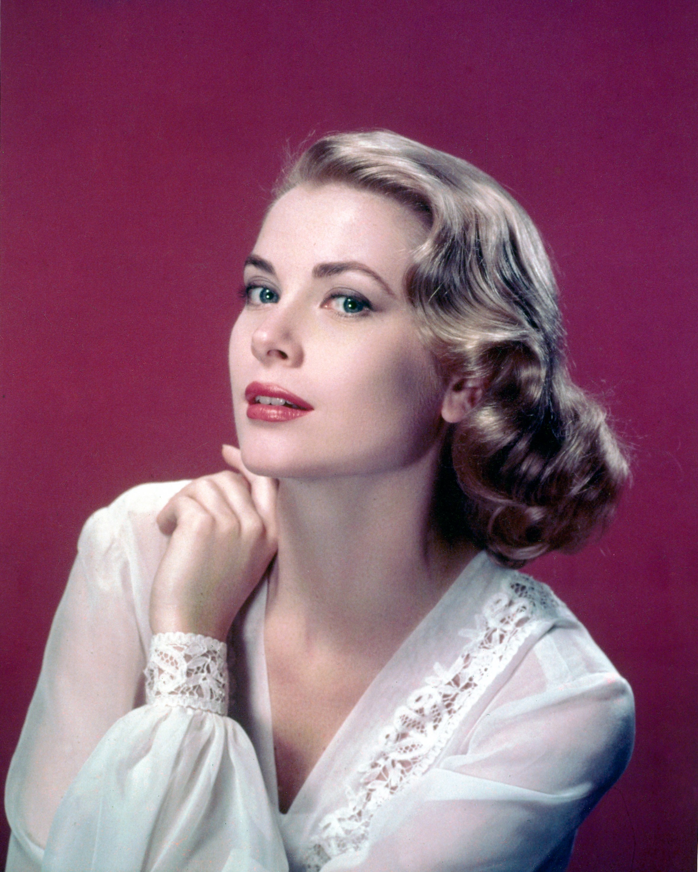 Grace Kelly posing against a red background, wearing a white blouse with lace trim.