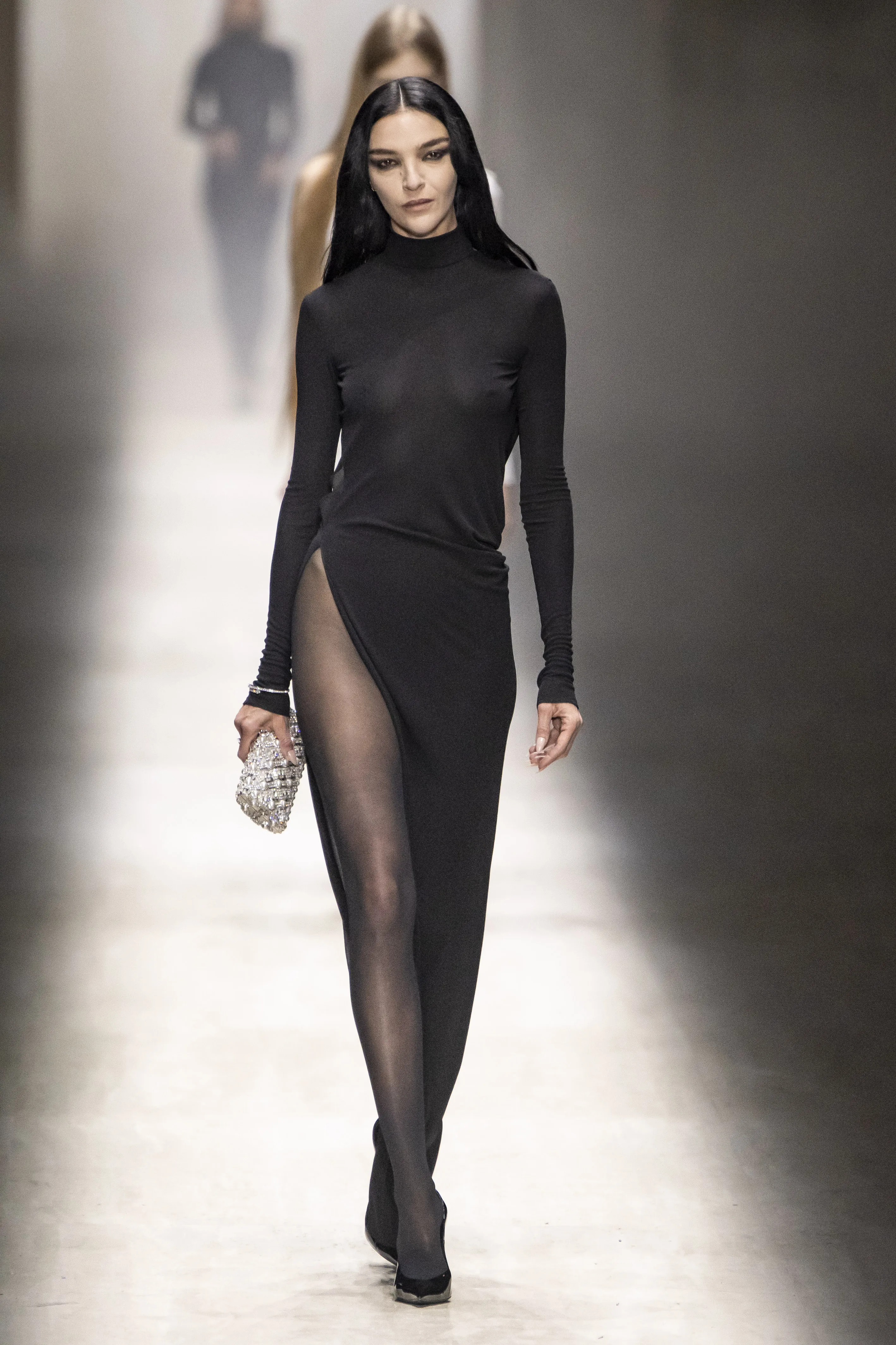 Mariacarla Boscono walking the runway in a sheer black turtleneck dress with a high slit, carrying a sparkling clutch.