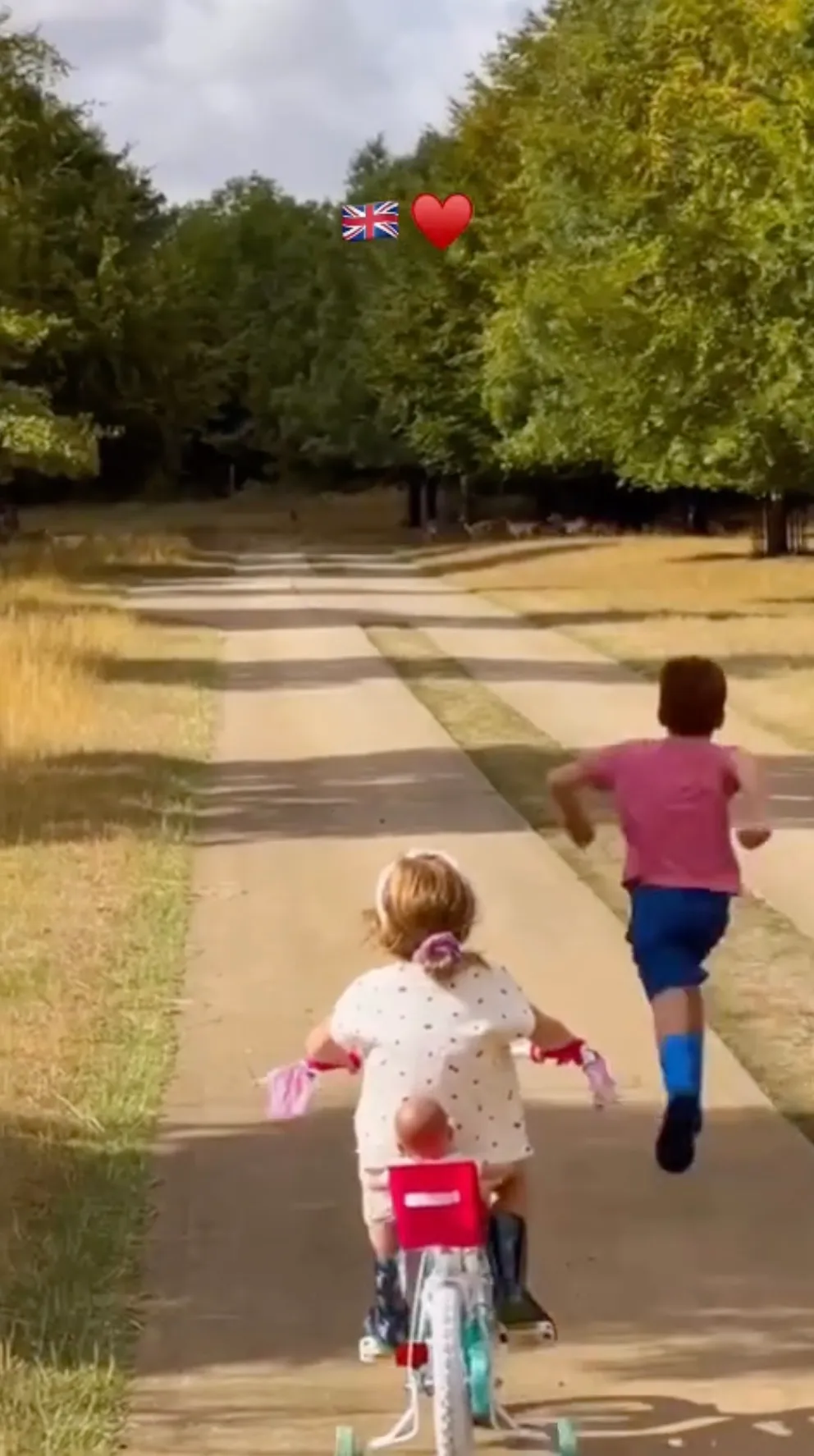 Two children, one riding a bicycle and one running, on a path surrounded by trees, with a Union Jack flag and red heart emoji overhead.
