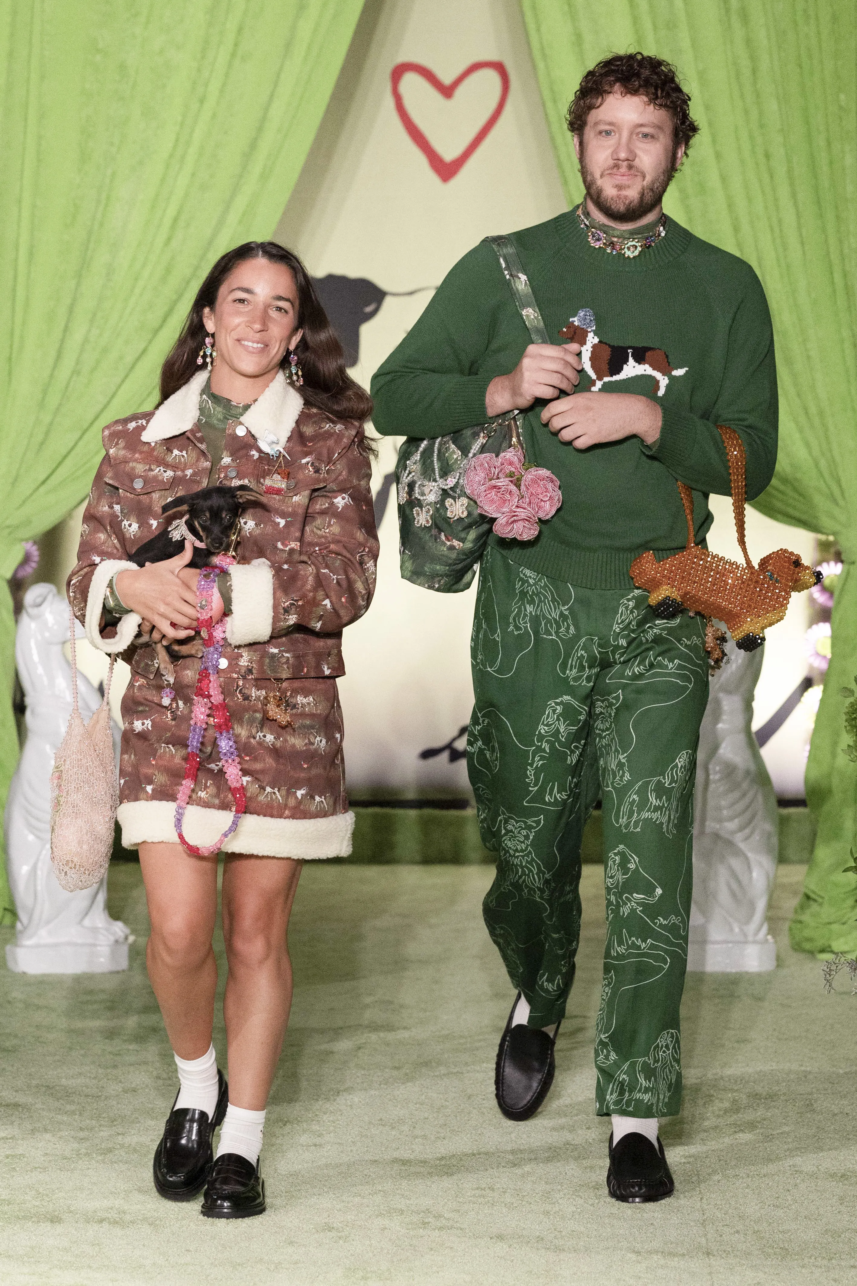 A woman holding a tiny puppy and a man wearing a sweater with a dog illustration walk down a runway.