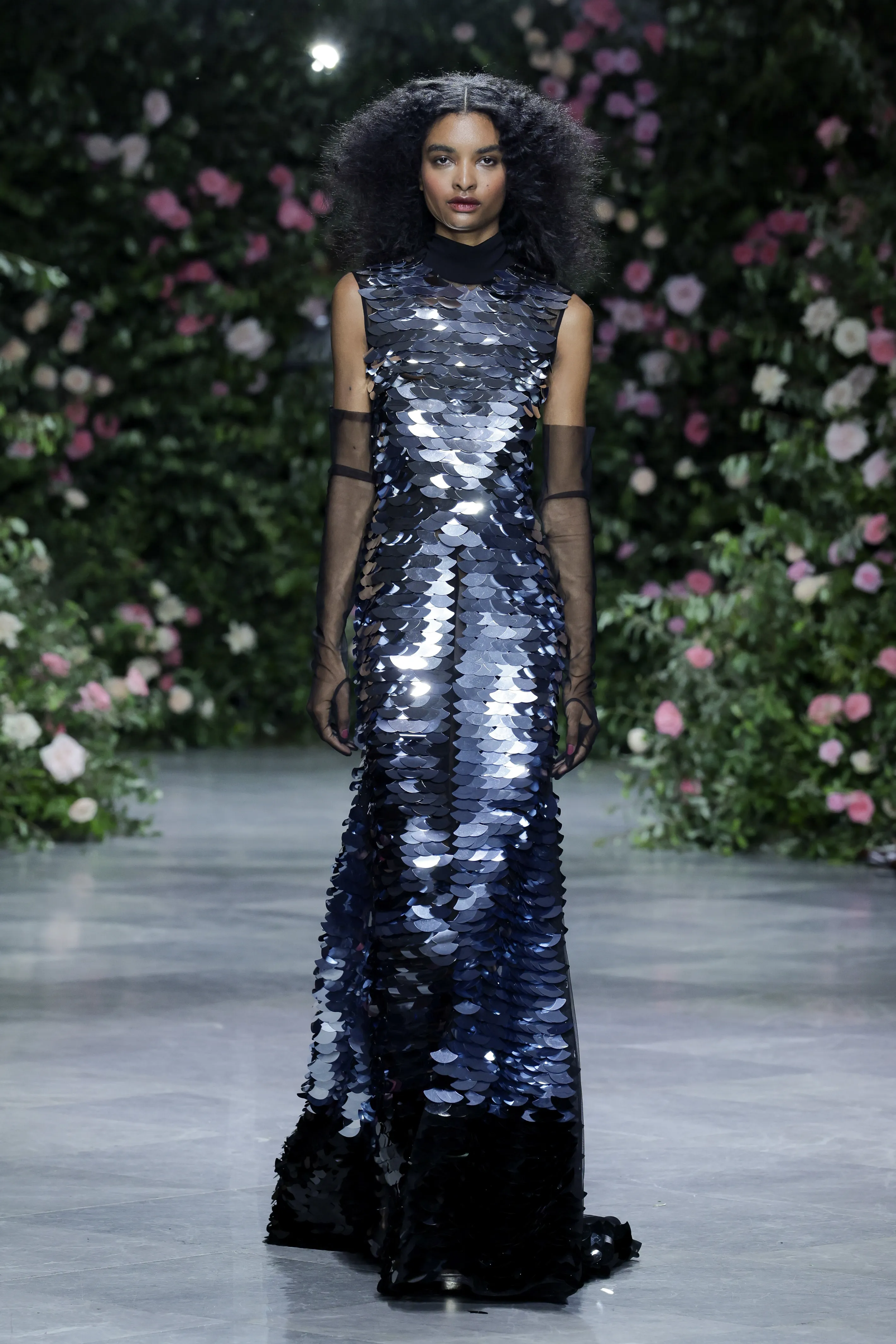 A model walks the runway in a long, sleeveless dress made of metallic sequins, with sheer black opera gloves.