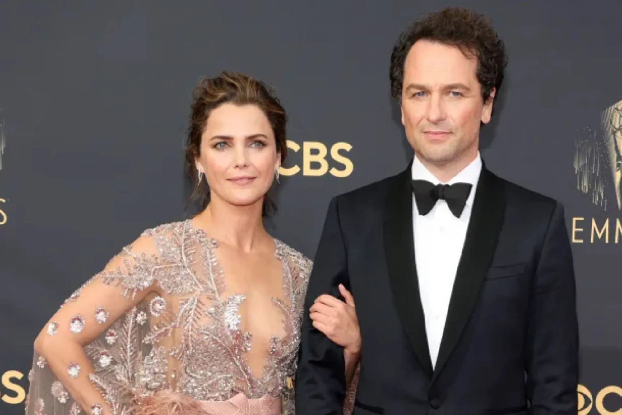 An image collage containing 1 images, Image 1 shows Keri Russell, In a metallic gown, and Matthew Rhys, in a black tuxedo, on the red carpett the Emmys