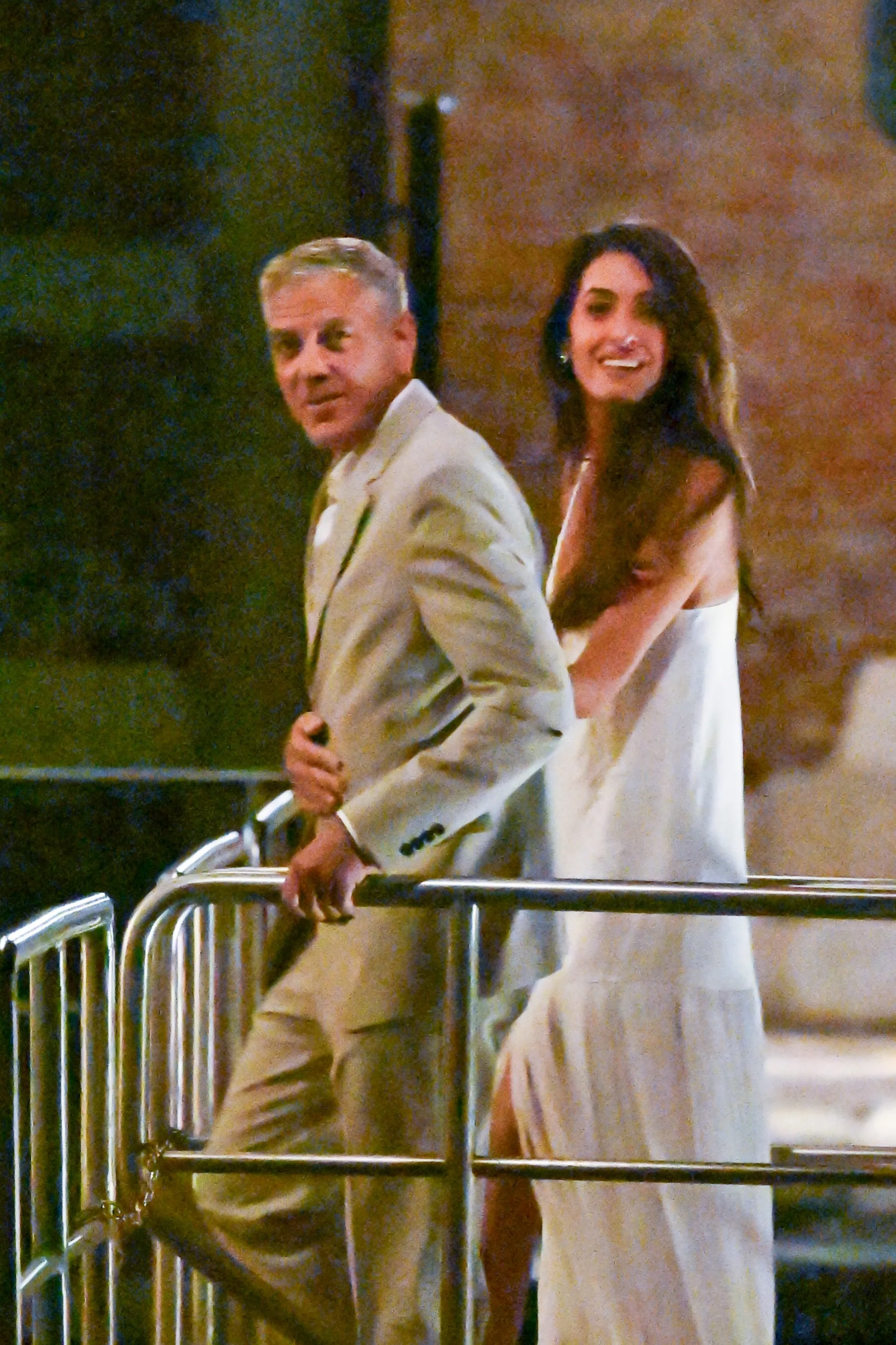 George and Amal Clooney smile in formal wear, George in a light gray suit and Amal in a flowing white gown, revisiting their wedding setting in Venice.